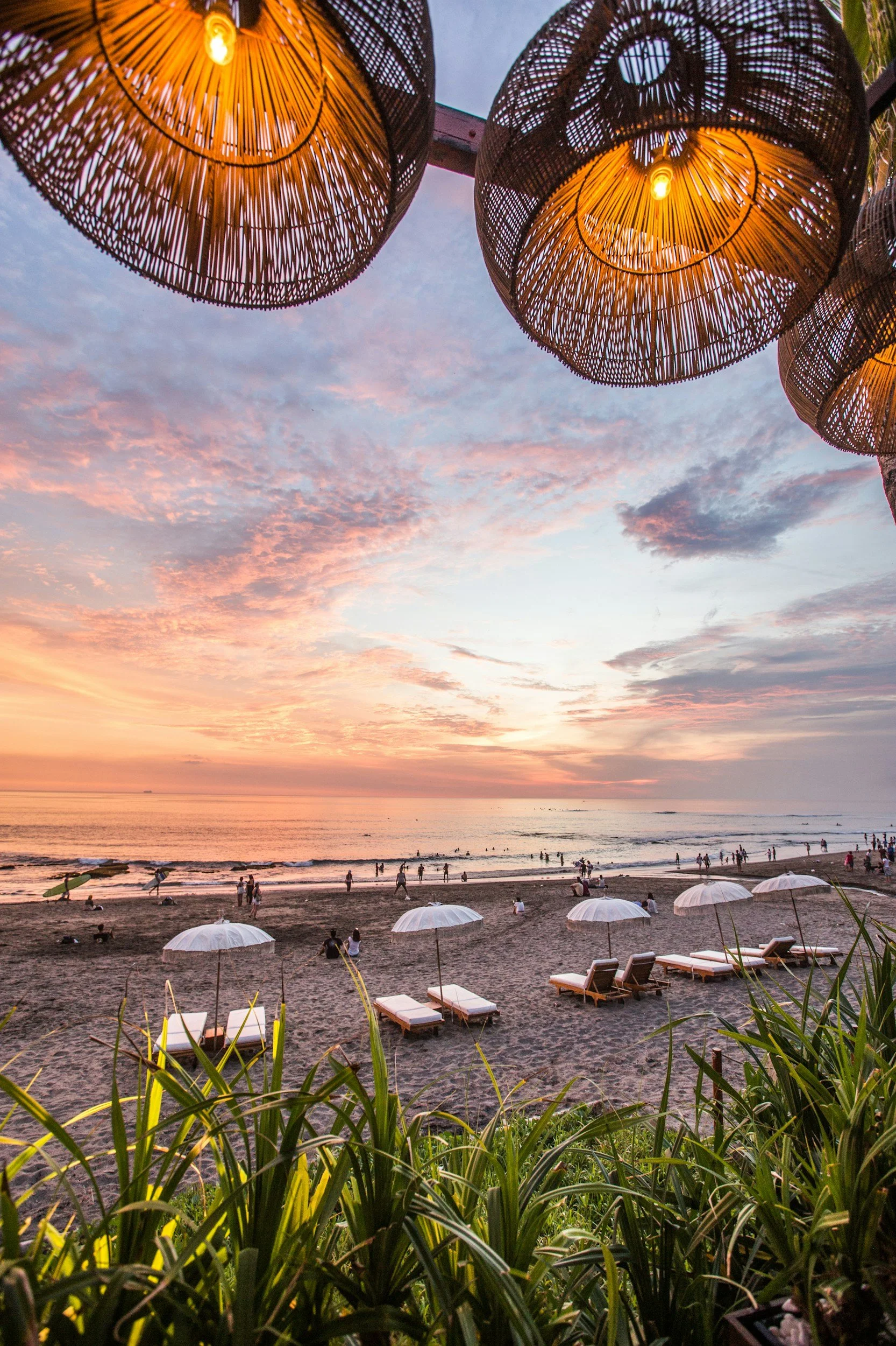 A beach scene at sunset with colorful clouds, lounge chairs, umbrellas, and people near the water, viewed from behind some green plants, with hanging woven lamps overhead.