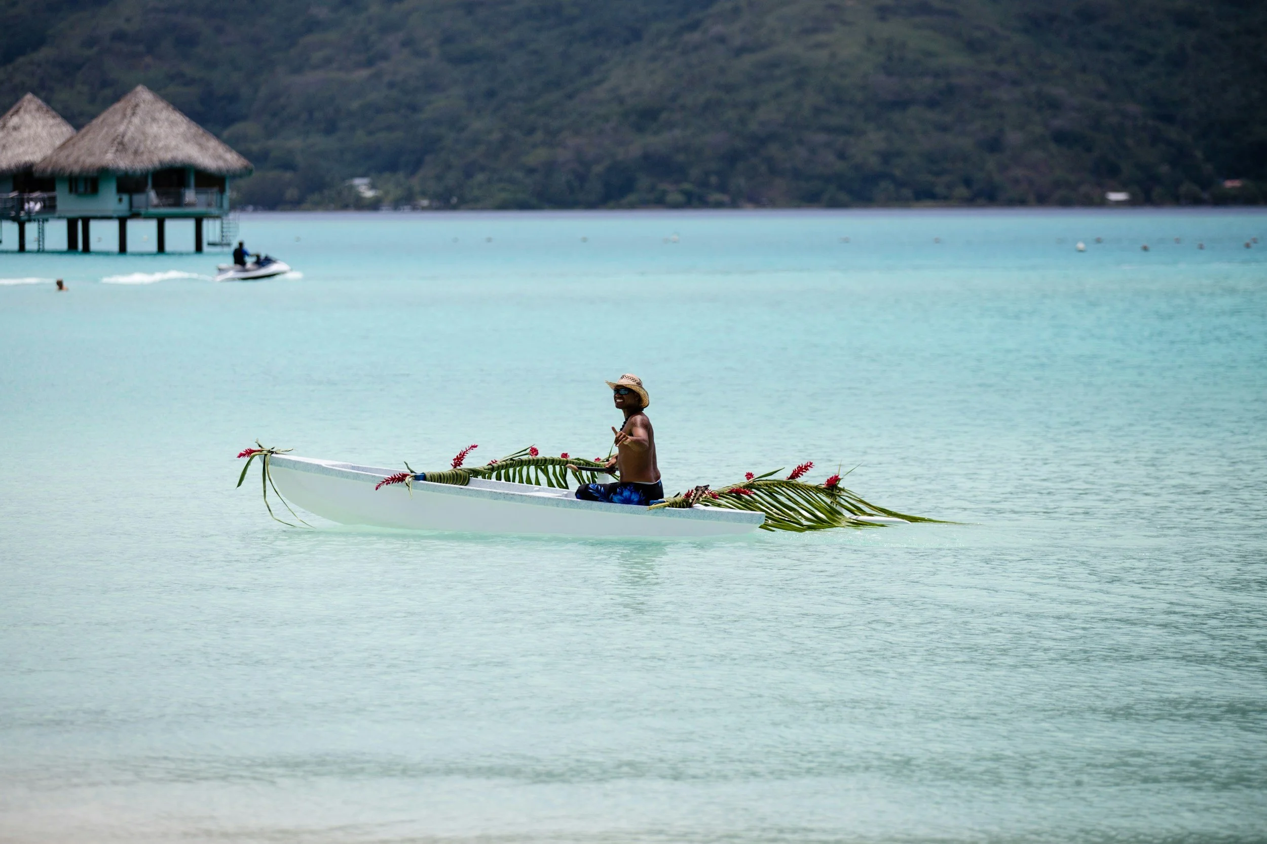 A man sitting in a canoe decorated with tropical leaves and flowers, floating on calm turquoise water near beach resorts with thatched roofs and a forested hillside in the background.