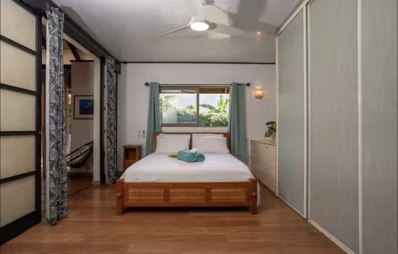 A cozy bedroom with a wooden bed frame, white bedding, and turquoise towels on the bed. There are curtains on a window behind the bed, a small white dresser on the right, and a hanging lamp on the wall. The room has wooden flooring and sliding closet