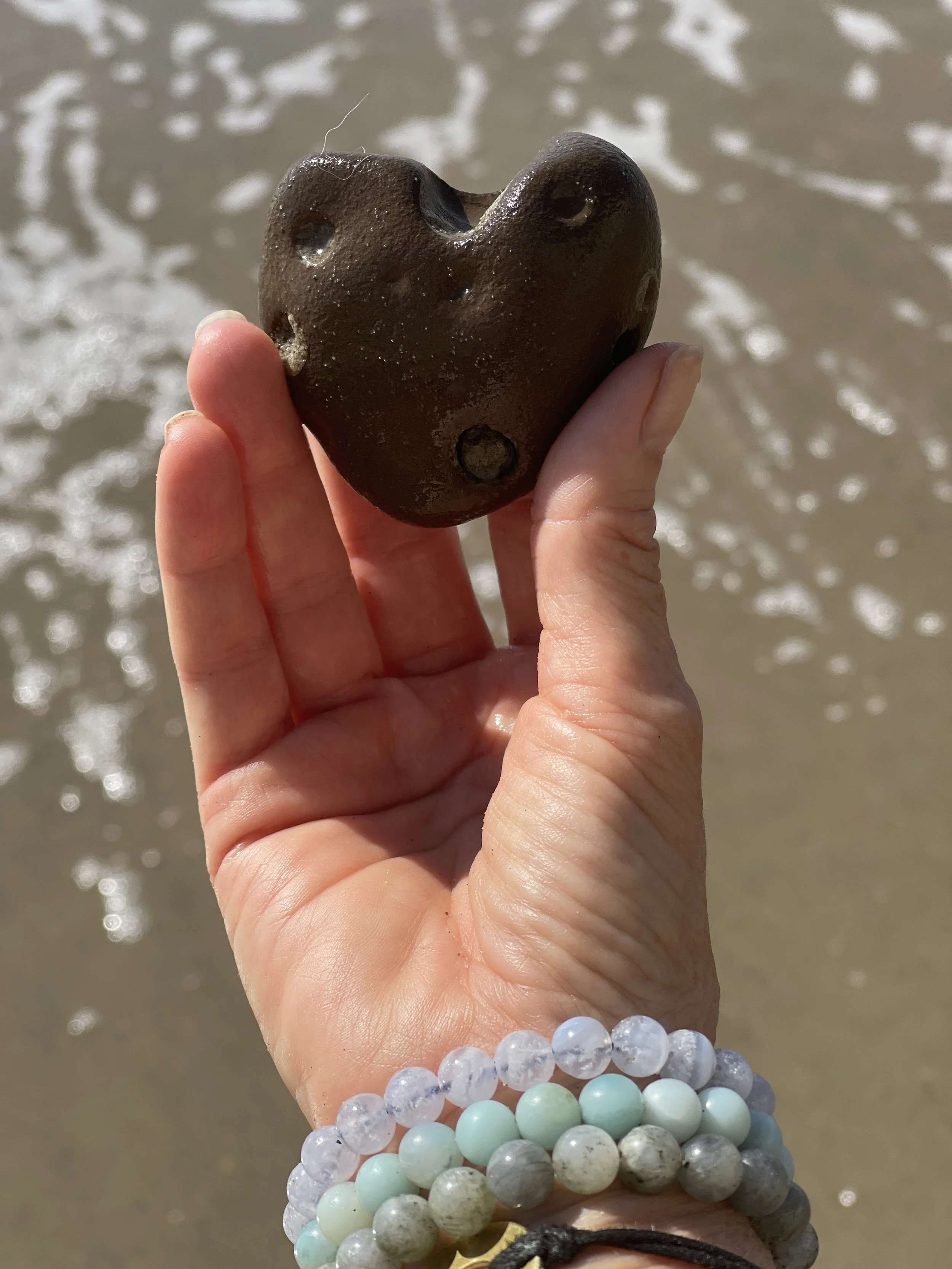 Person holding a dark, heart-shaped rock with holes near the beach, with water and waves in the background.