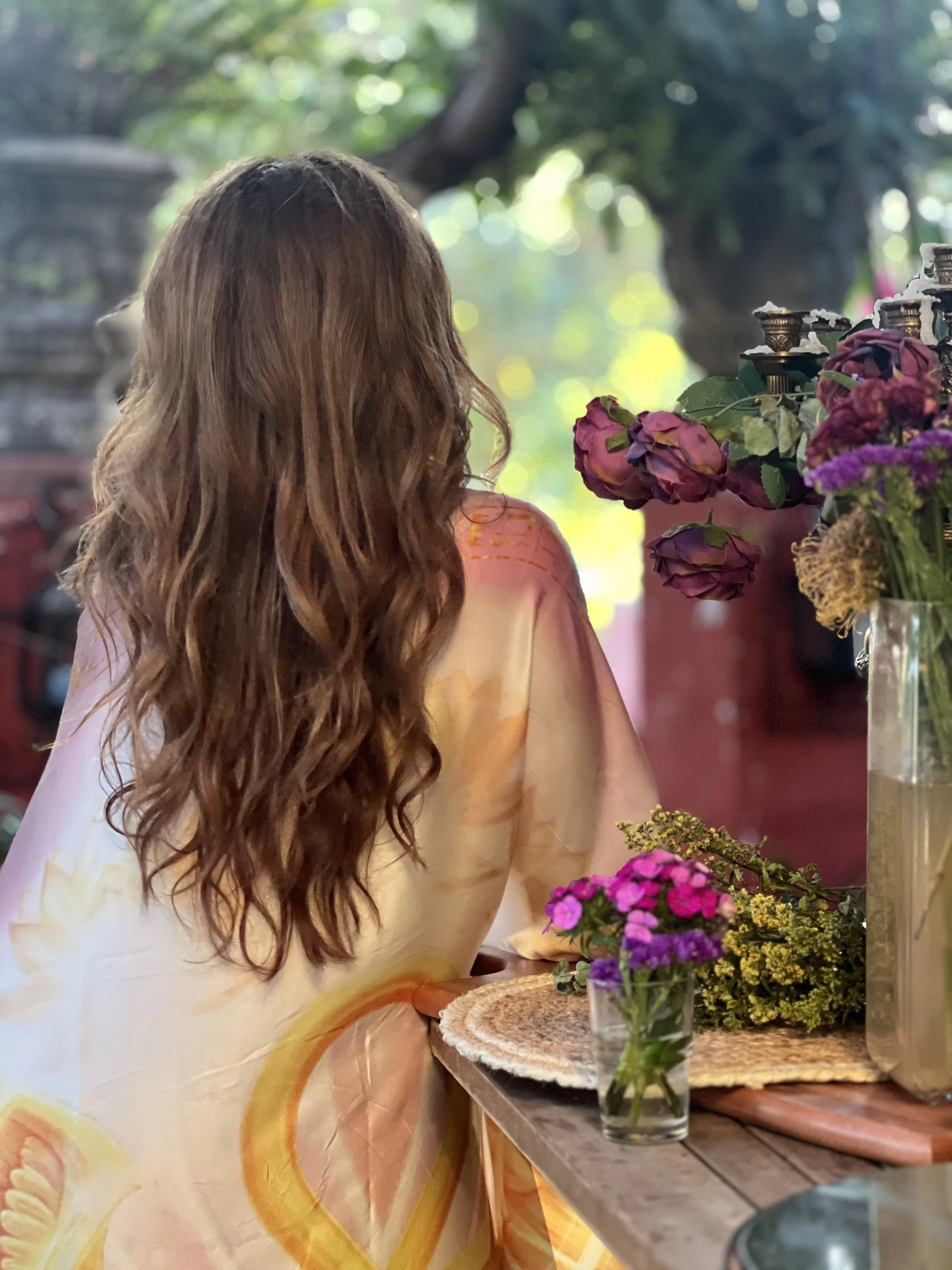A woman with long, wavy hair viewed from the back, sitting at a table with floral arrangements in a cozy, outdoor setting.