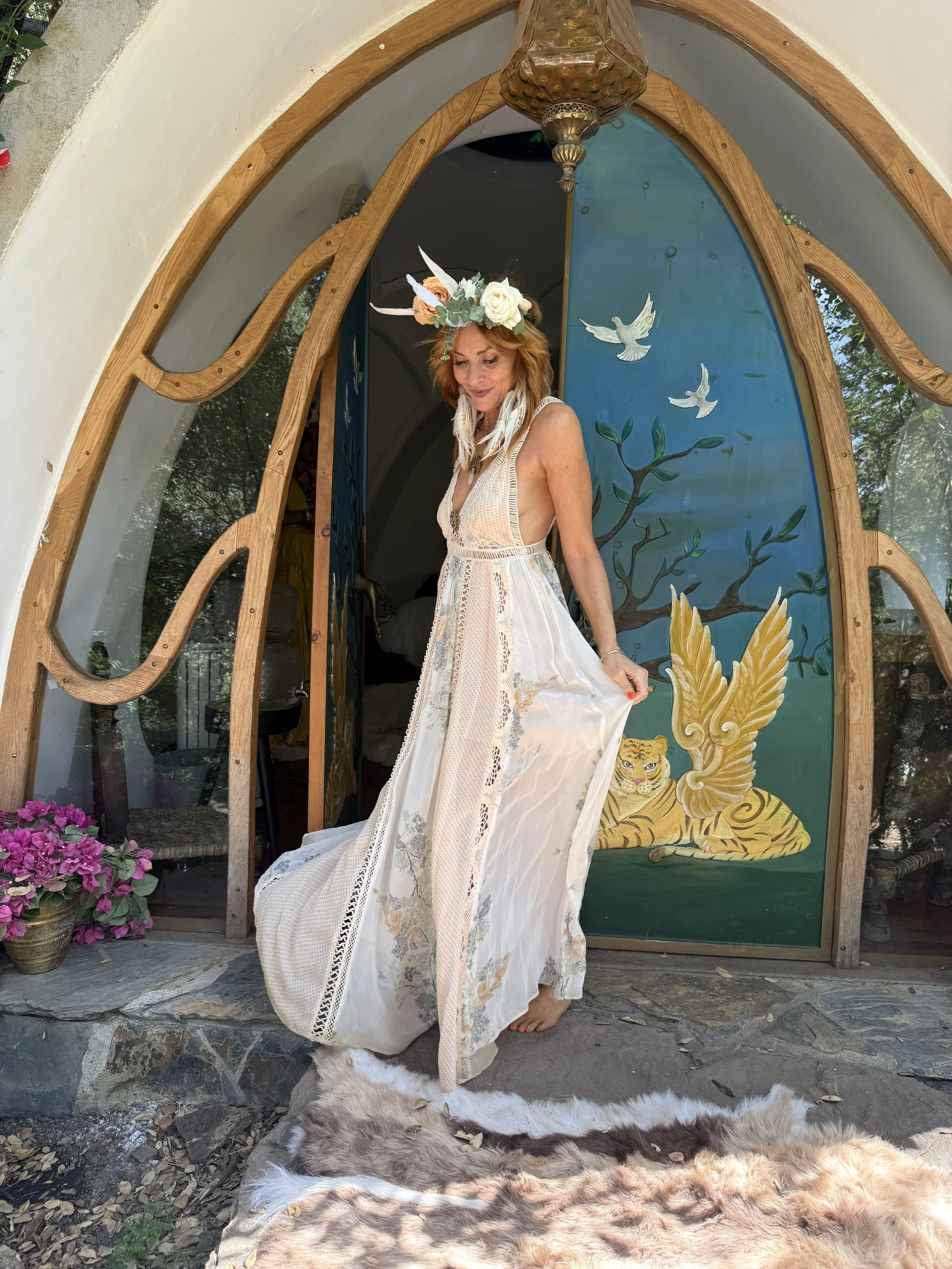 Woman dressed in a bohemian style white dress with lace details, barefoot, standing in front of a colorful painted door featuring a tiger, branches, and flying doves. She wears a flower and feather headpiece with horns, and holds the hem of her dress