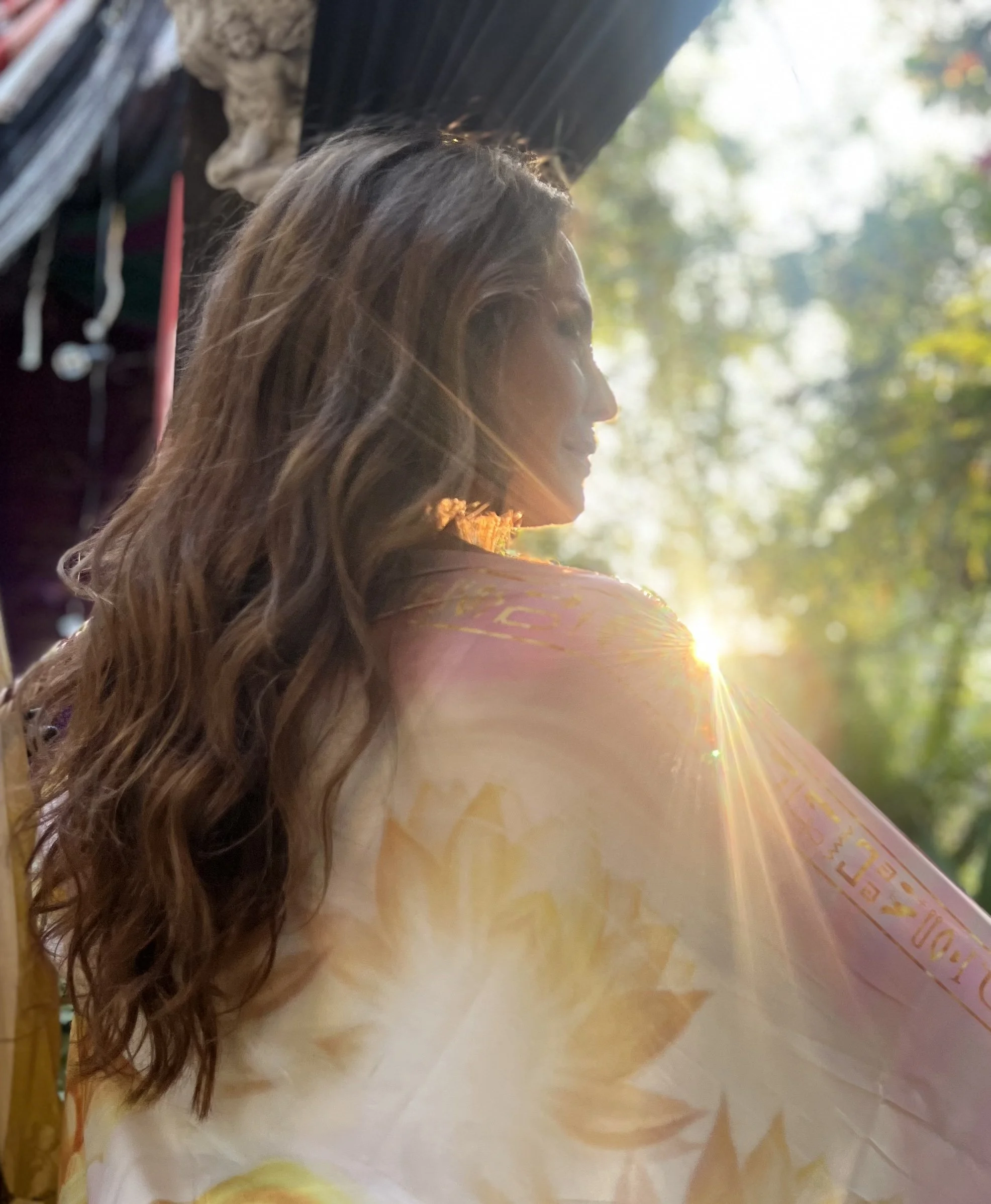A woman with long, wavy hair standing outdoors in sunlight, with trees in the background and sunlight creating a lens flare.