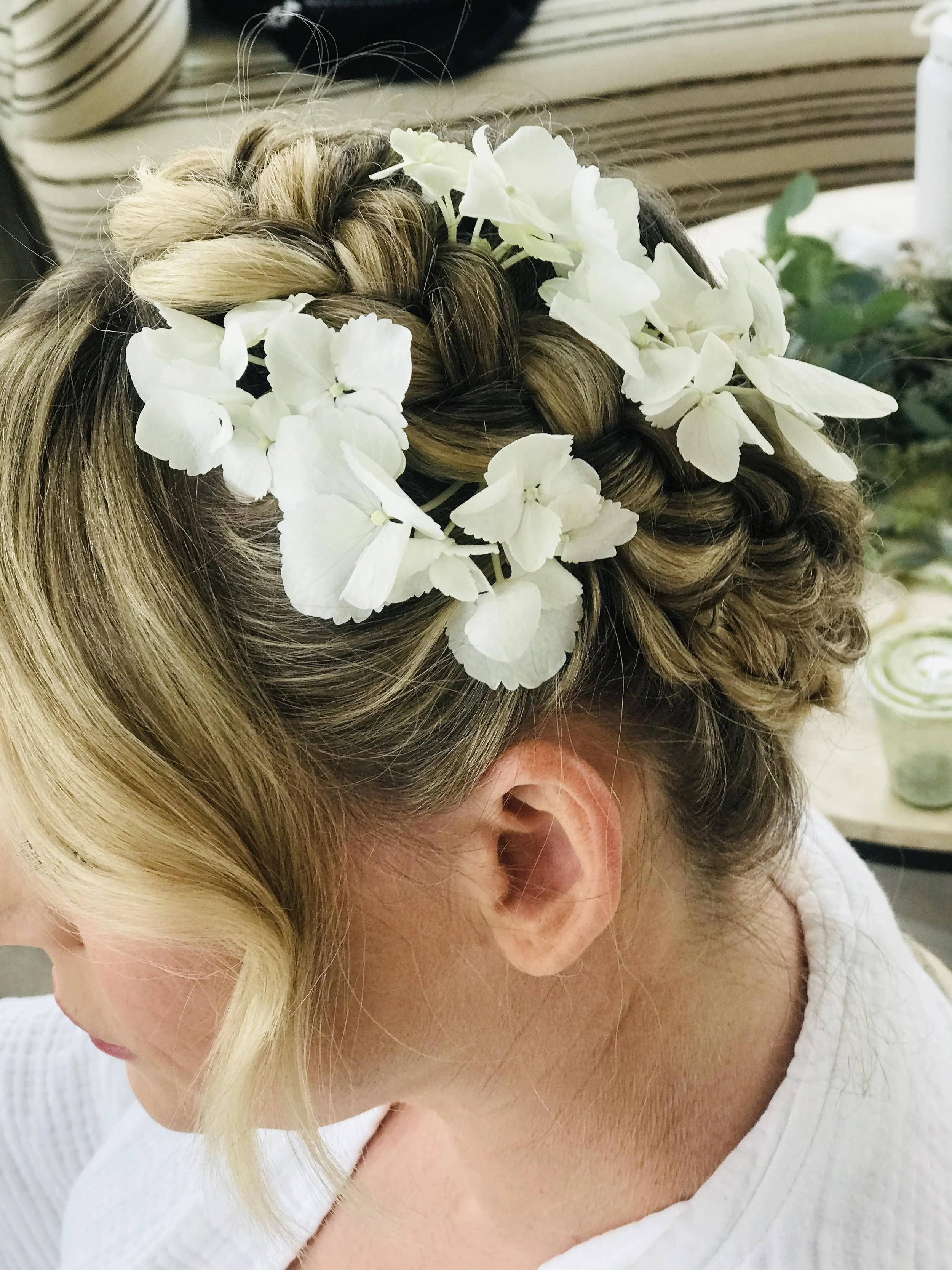 Close-up of a woman's head with intricately braided hair adorned with white hydrangea flowers, wearing a white top.