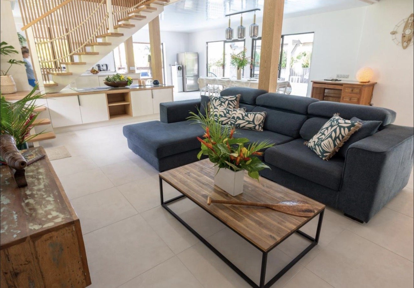 Living room with a dark blue sectional sofa with patterned pillows, a wooden coffee table with a flower vase, a modern kitchen with white cabinets, a staircase, and large windows letting in natural light.