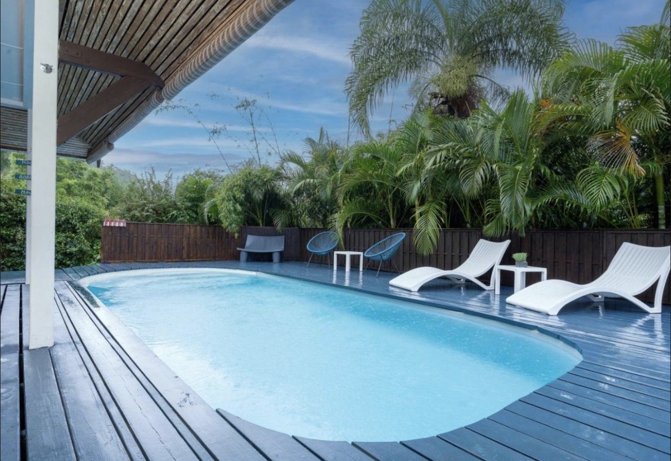 Outdoor swimming pool with a blue deck, surrounded by tropical plants and white lounge chairs, under a partly cloudy sky.