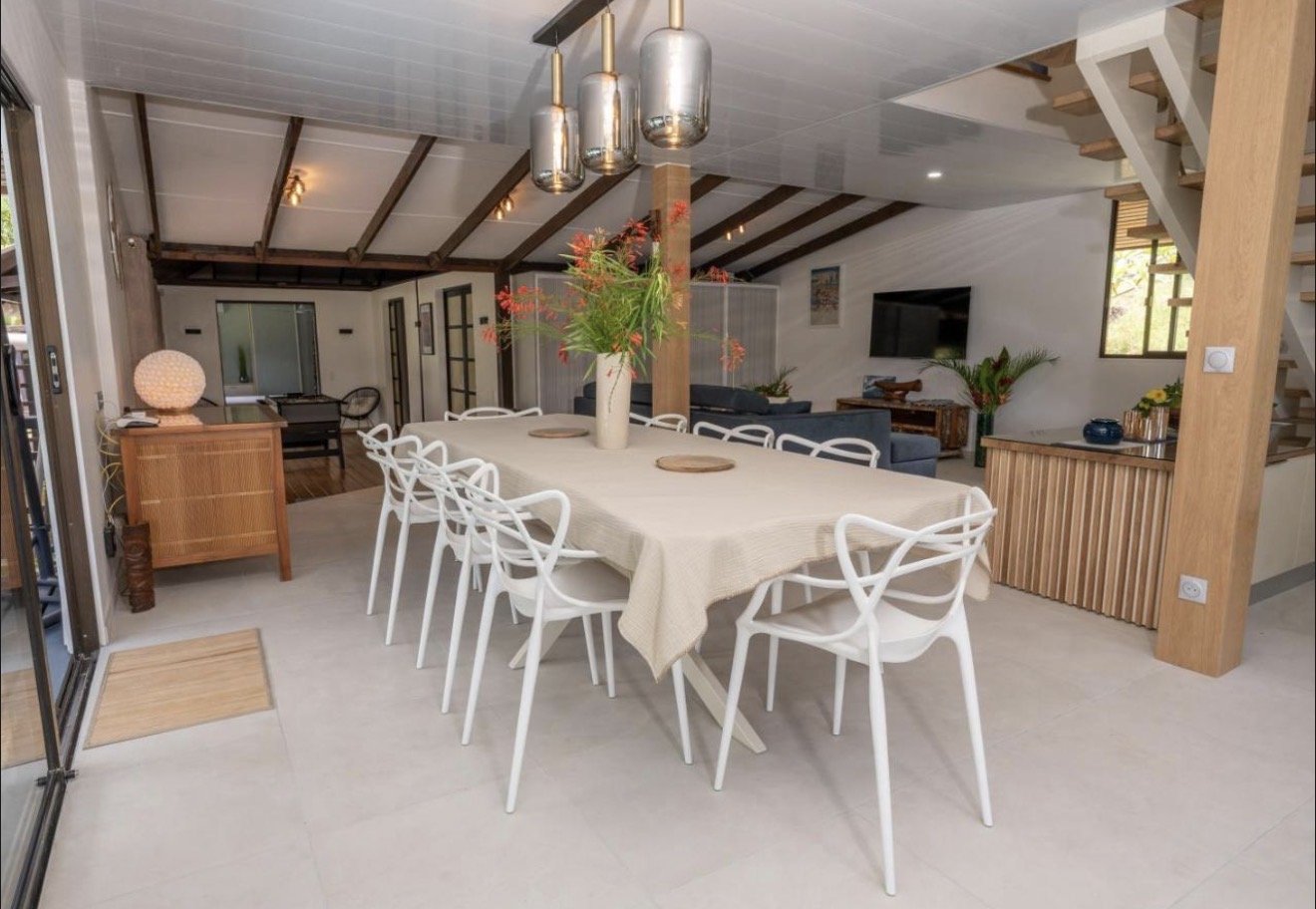 Interior view of a dining area with a long beige table, white chairs, and hanging pendant lights. The room has a sloped ceiling with wooden beams and a staircase in the corner. There is a vase with flowers on the table and various decorative plants i
