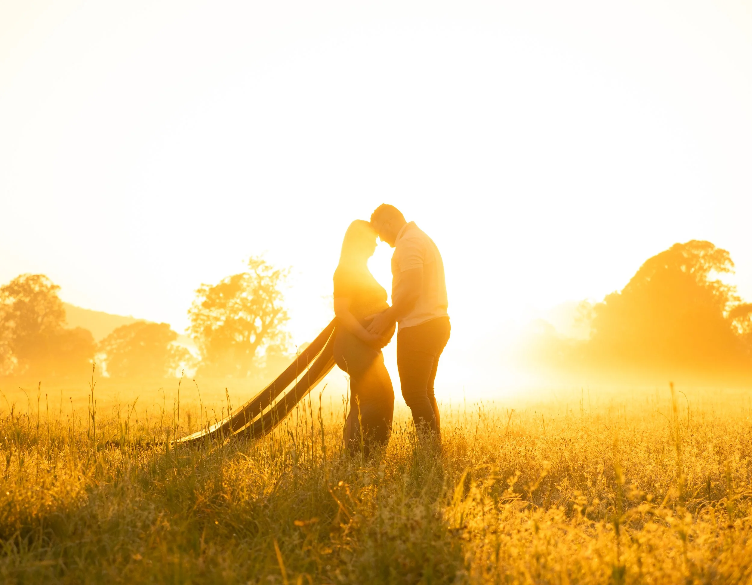 A couple embracing with the man's hands on his wife's preganant belly in the golden mornign light of a maternity session in the open fields of the Scenic Rim.