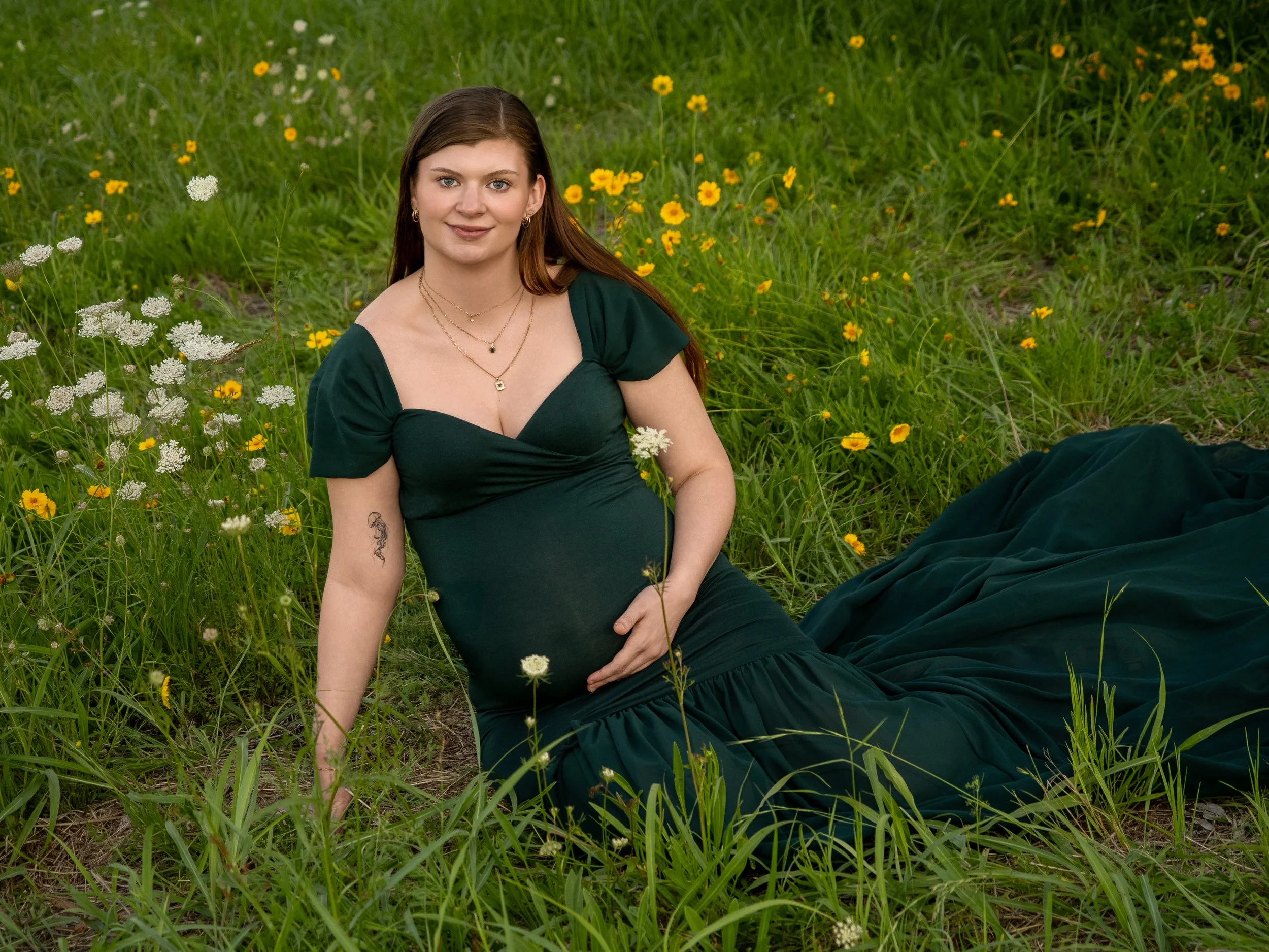 Expectant mother in a deep green dress holding her belly as she sits on the flower fields of Ipswich during a maternity session.