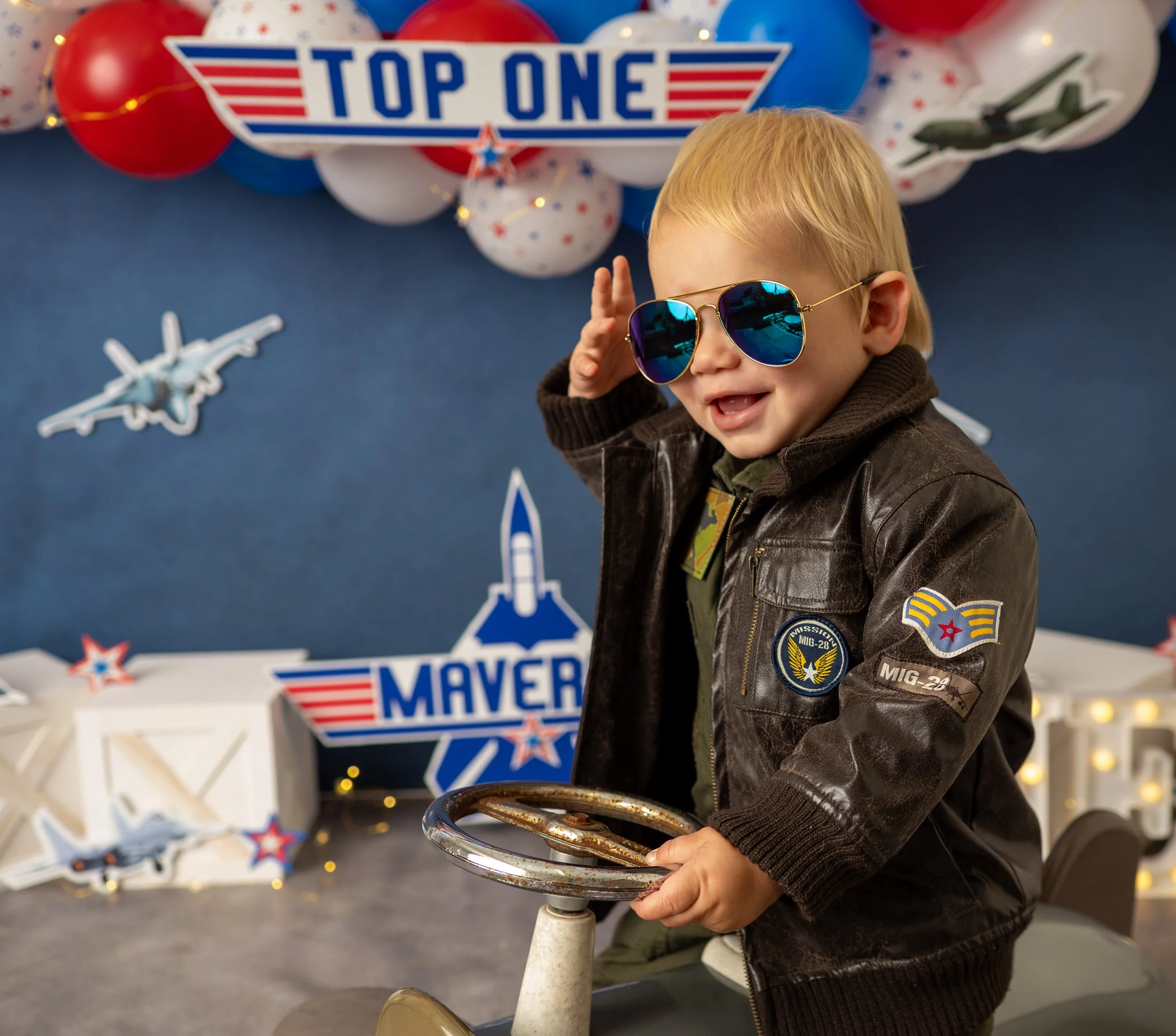 Little boy woith blue aviator sunglasses and a flight jacket on a ride-on place for his first birthday portraits in a studio setting with the "Top One" cake smash theme.