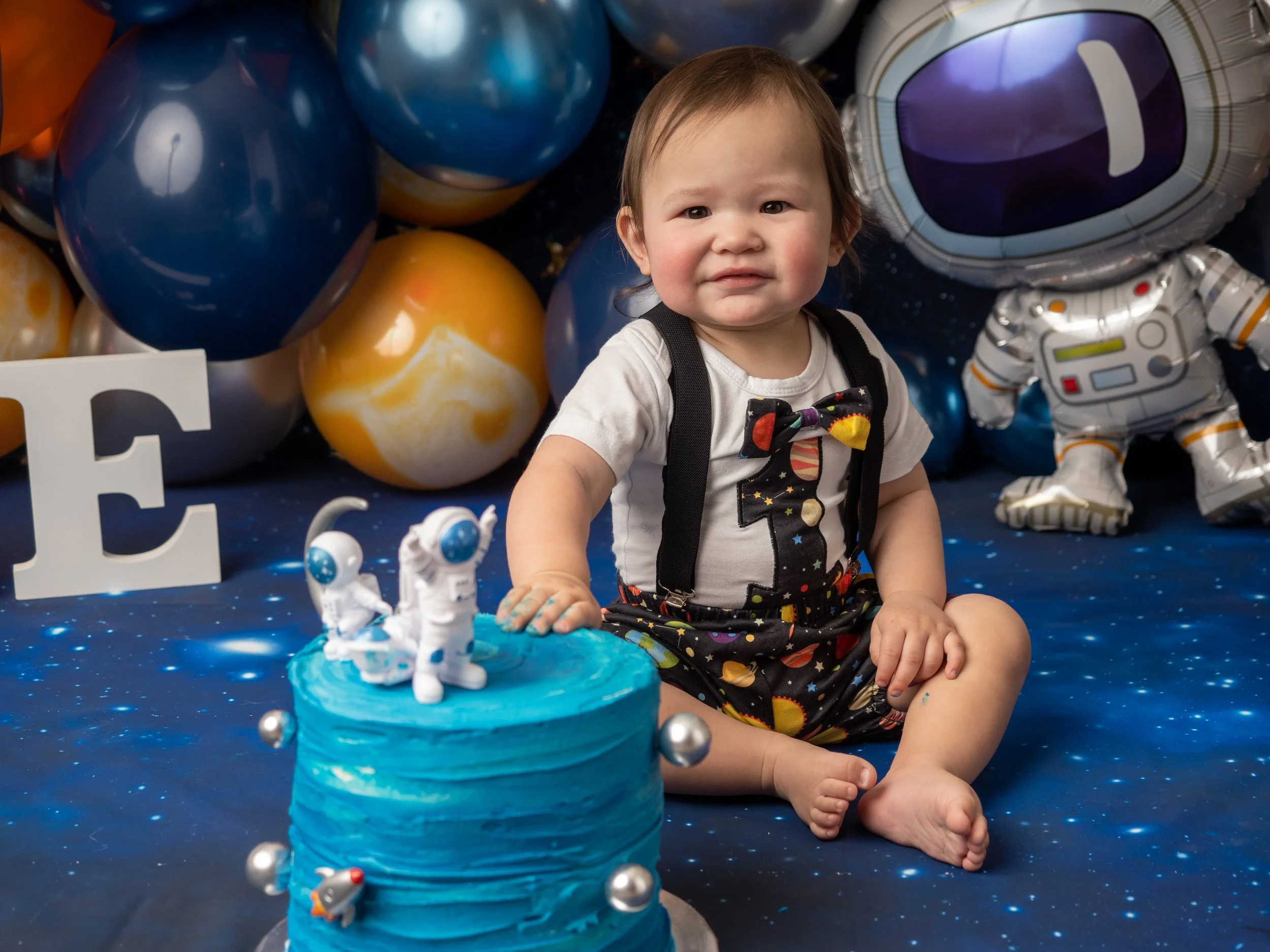 a little boy in a space themed romper with a 1 on it infront of a blue astronaut cake with balloons and a space themed backdrops with stars for a space theed cake smash in a studio setting.