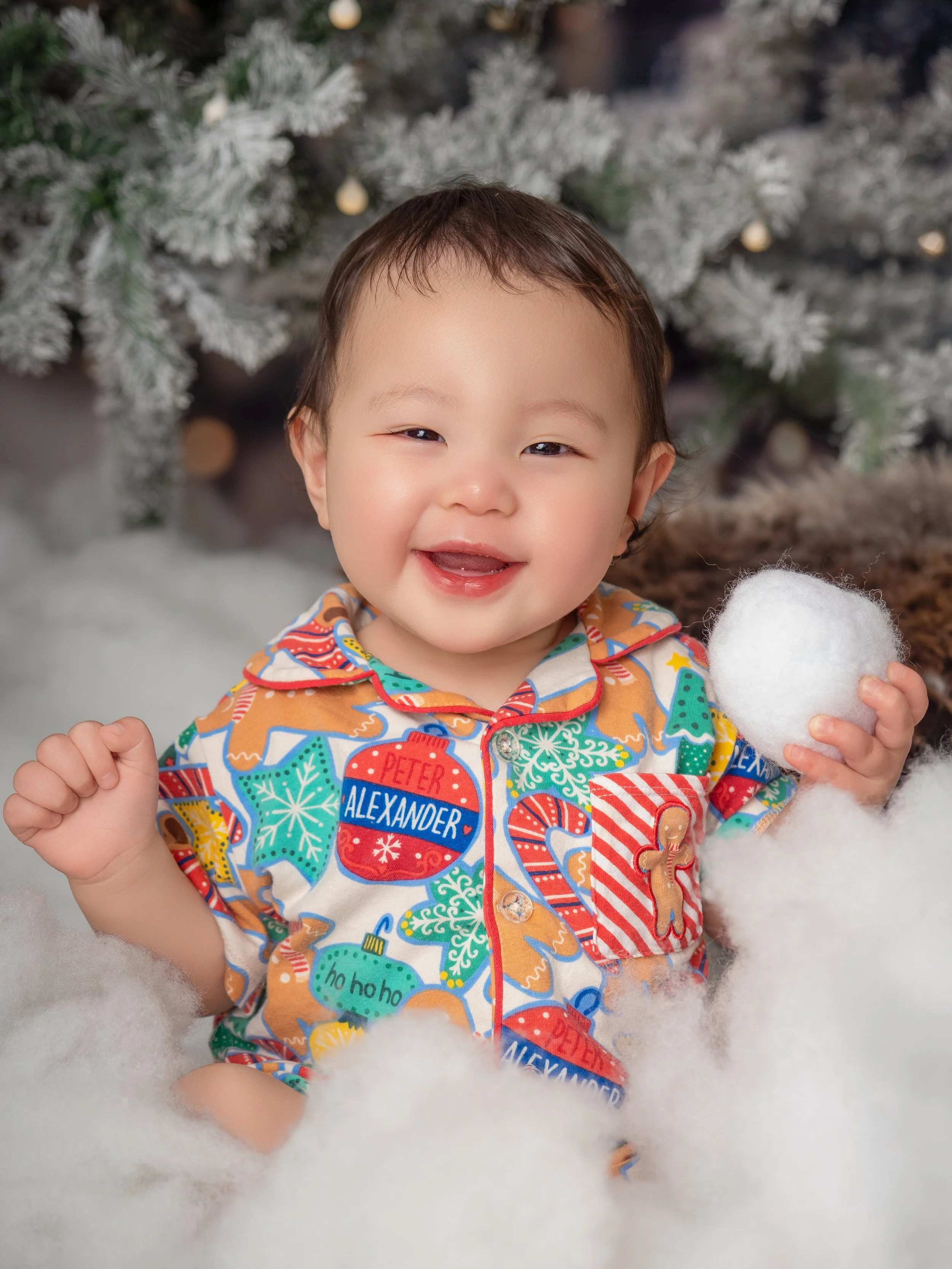 Young Asian baby boy in Christmas pajamas holding a snow ball and laughing enjoying the snowy Winter Wonderland Christmas studio session