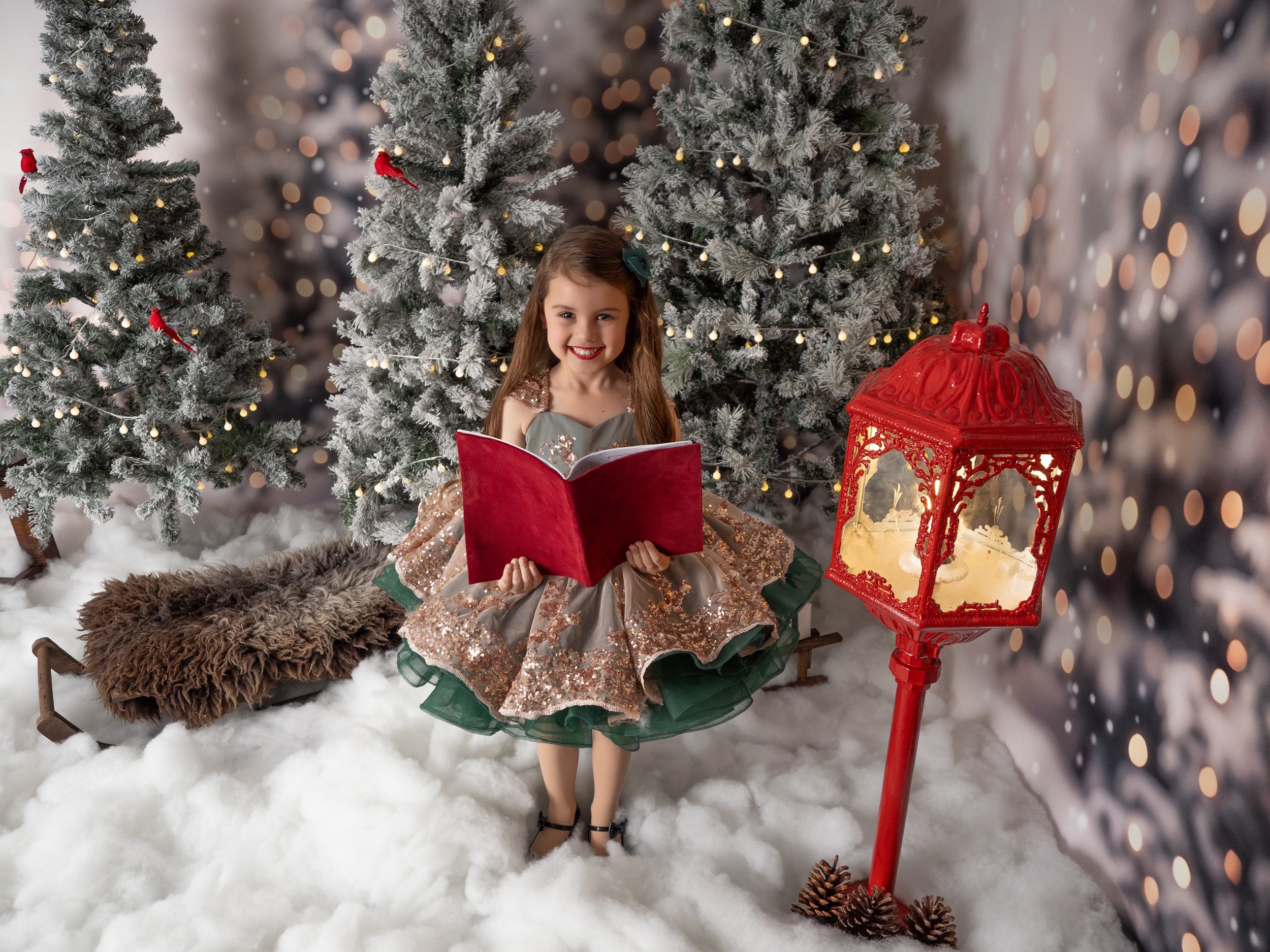 Young girl enjoying a Christmas carolling book surrounded by snowy Christmas trees at a Winter Wonderland mini session