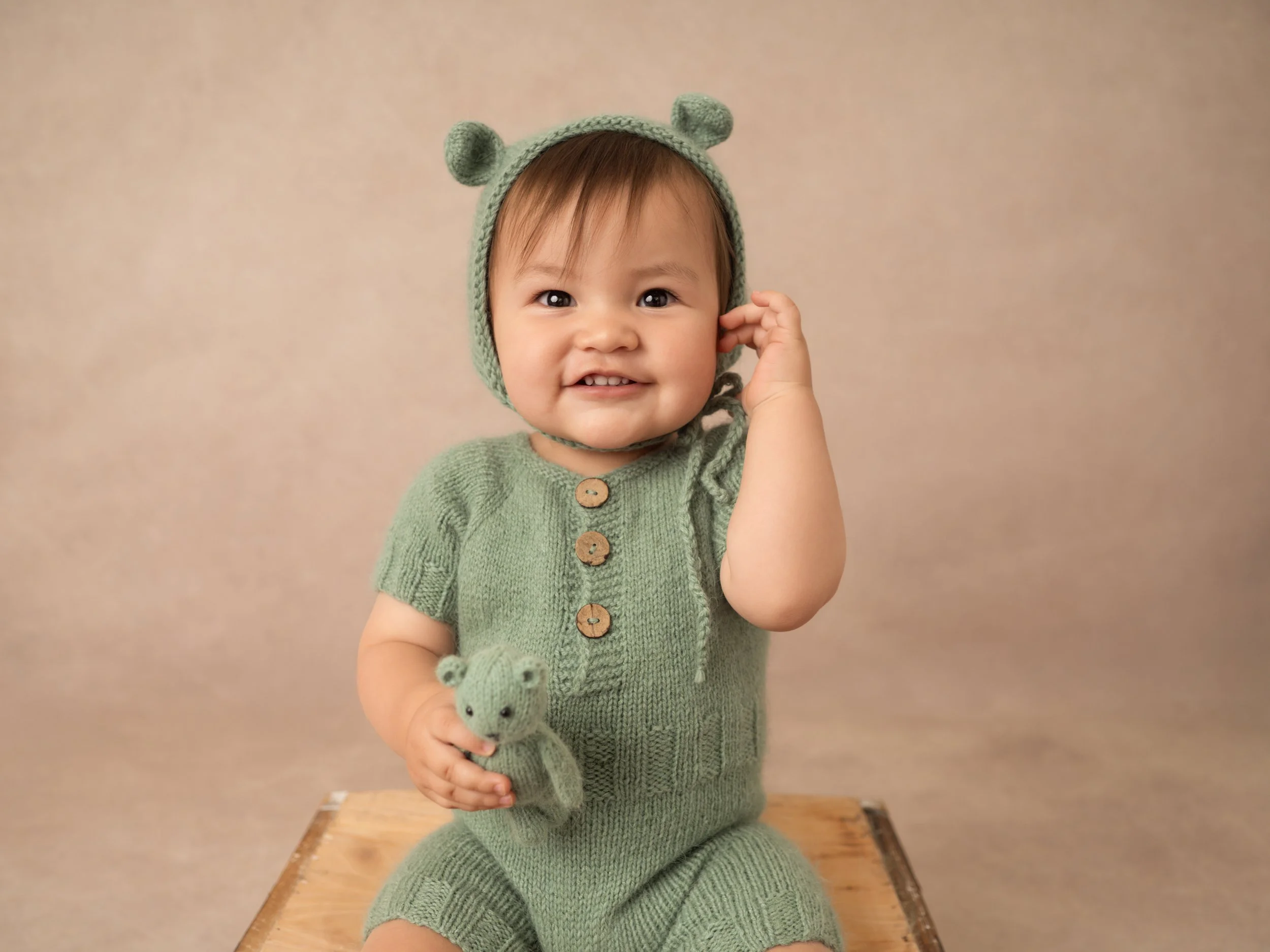 A little boy under 1year old in a little green knit outfit with knitted bear ear bonnet, holding a littel green knit teddy, as he smiles for a milestone studio potrait.
