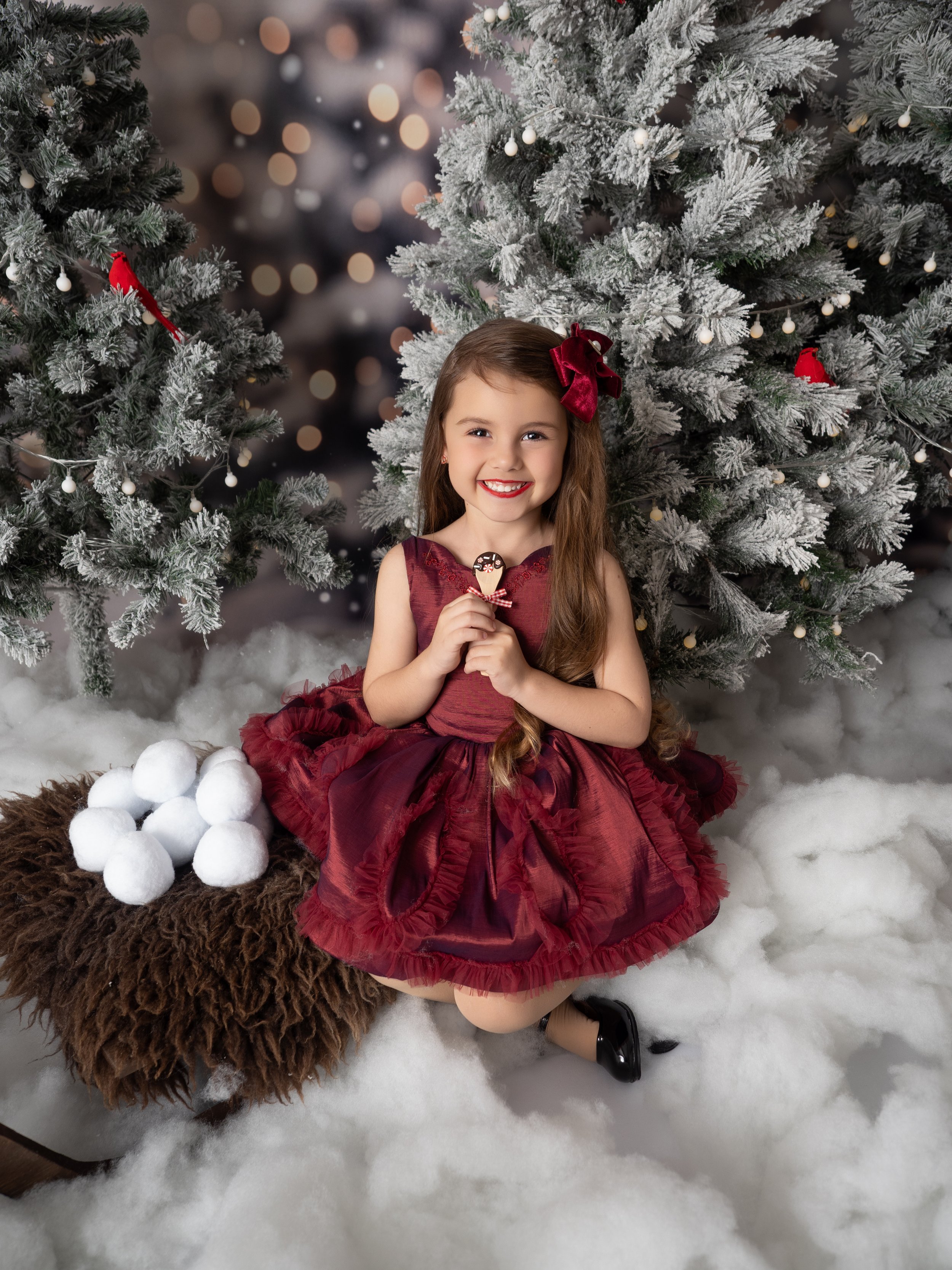 Young girl enjoying a Christmas chocolate sppon sitting on a sled with wooly layer and snow balls, surrounded by snowy Christmas trees at a Winter Wonderland mini session