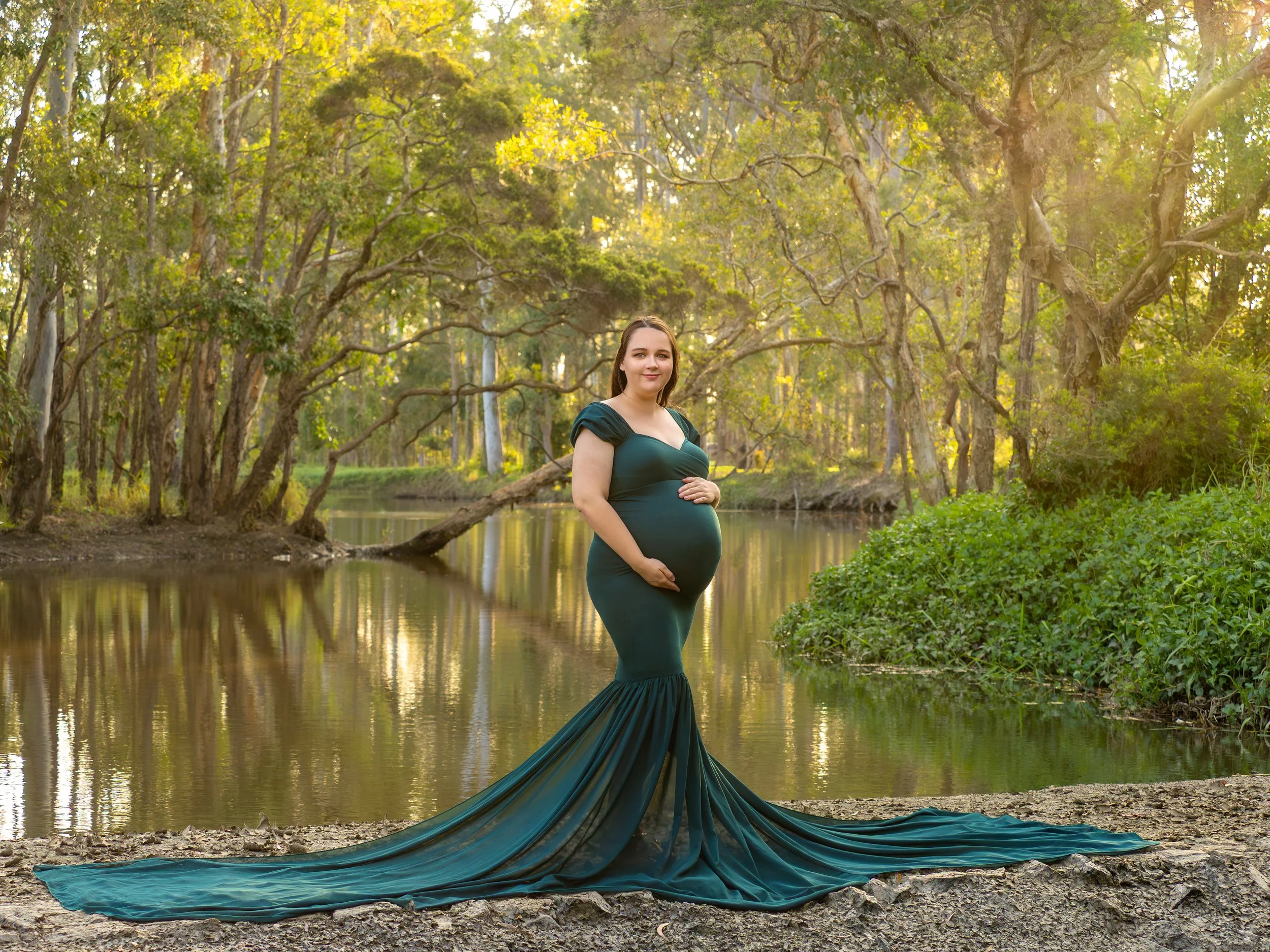 A pregantn lady holding her belly as she wear a forest green fitted dress with mermaid chiffon train, standing by a reiver bank in Brisbane for a golden hour maternity session.