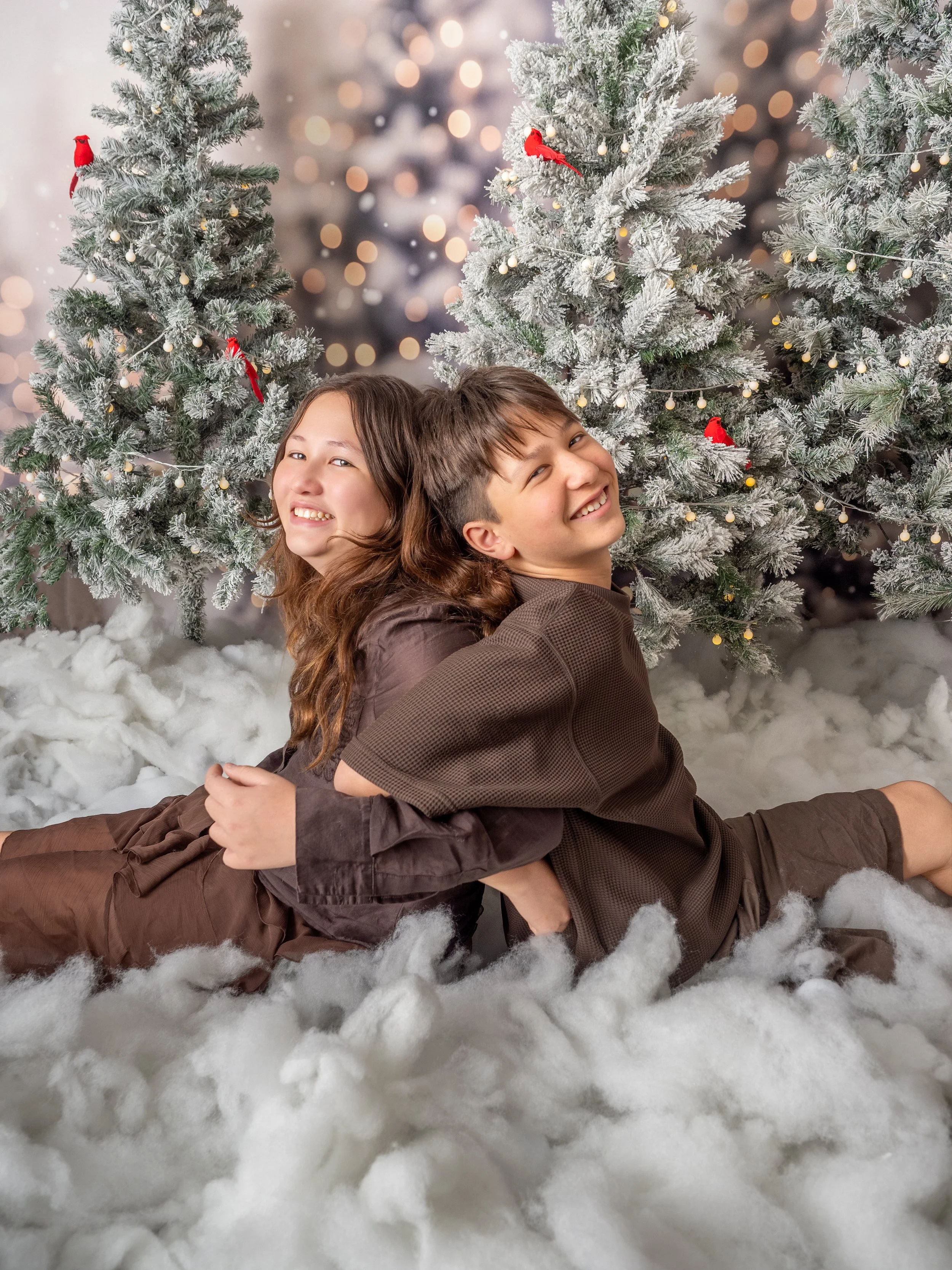 A pair of mixed siblings sitting back to back in hte snow laughing together surrounded by fairy lit snowy Christmas trees in the winter wonderland Christmas mini set