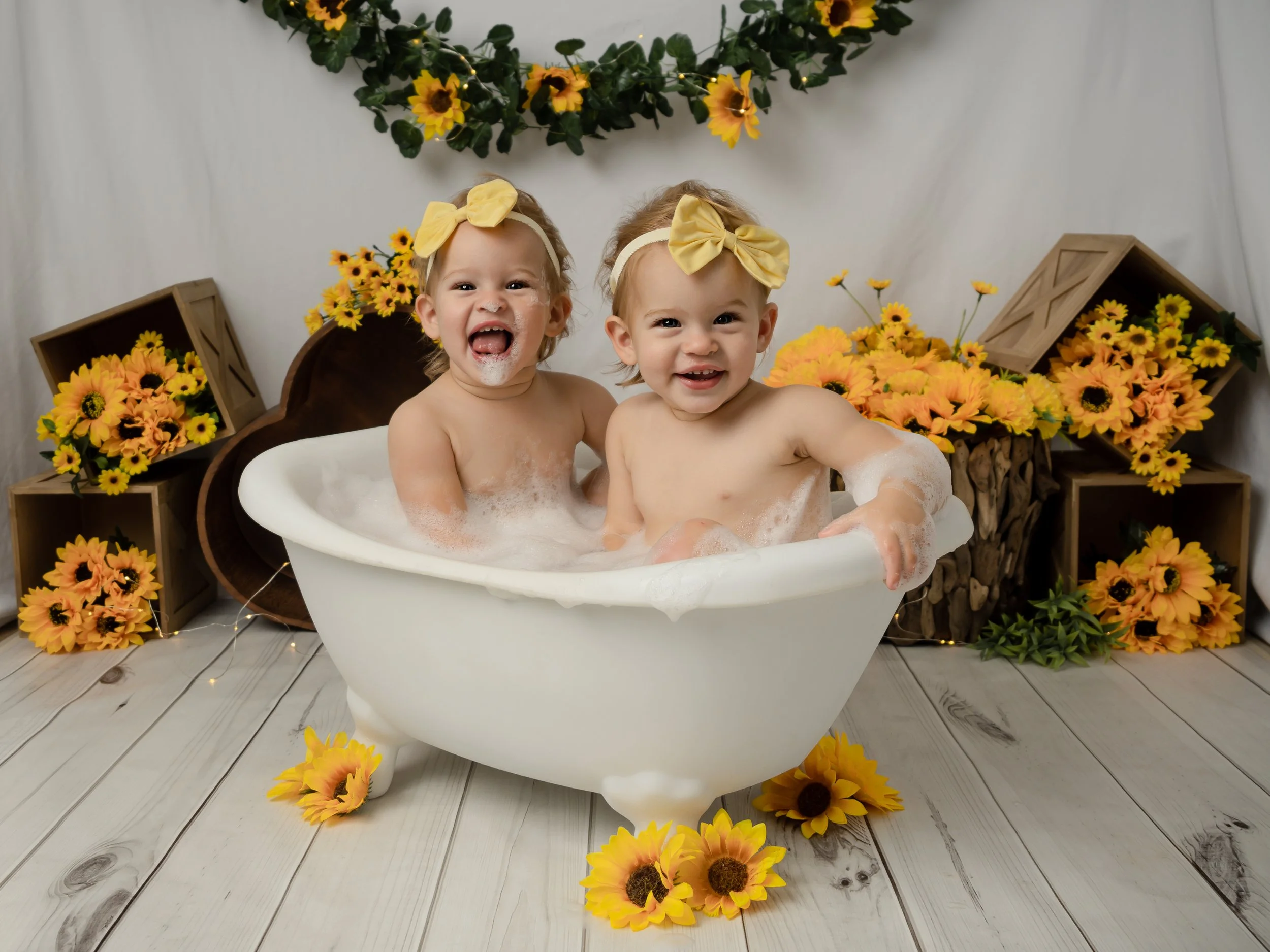 Identical twin girls with yellow bows in their hair splashing around in a small white bath tub filled with bubbles in a studio setting decorated with sunflowers for their 1st birthaday cake smash.