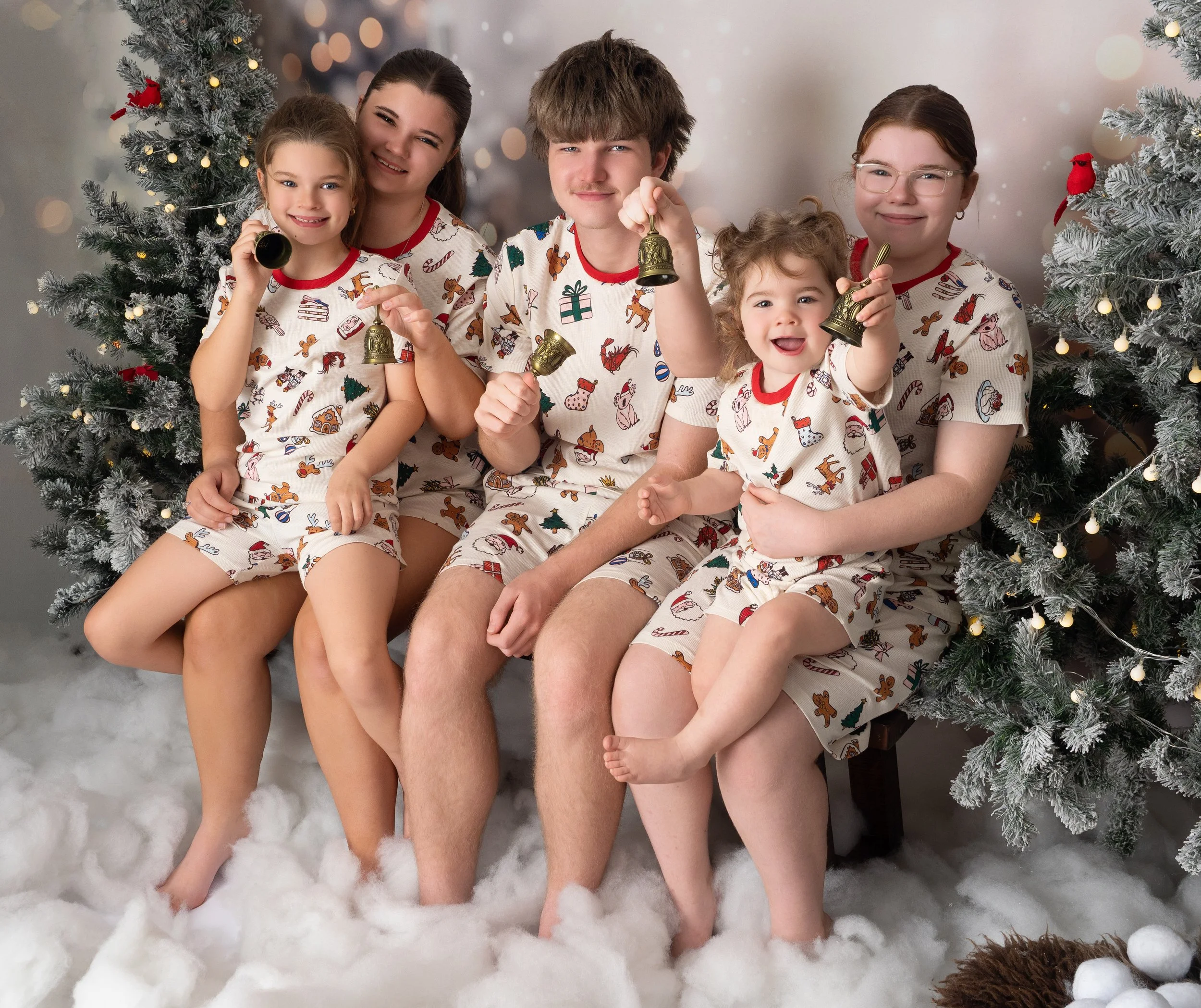 Five siblings in matching Christmas pajamas all playfully ringing small Christmas bells seated on a rustic wooden beanch surrounded byt snowy Christmast trees lit up with small bulb fairy lights as part of a studio Christmas mini set