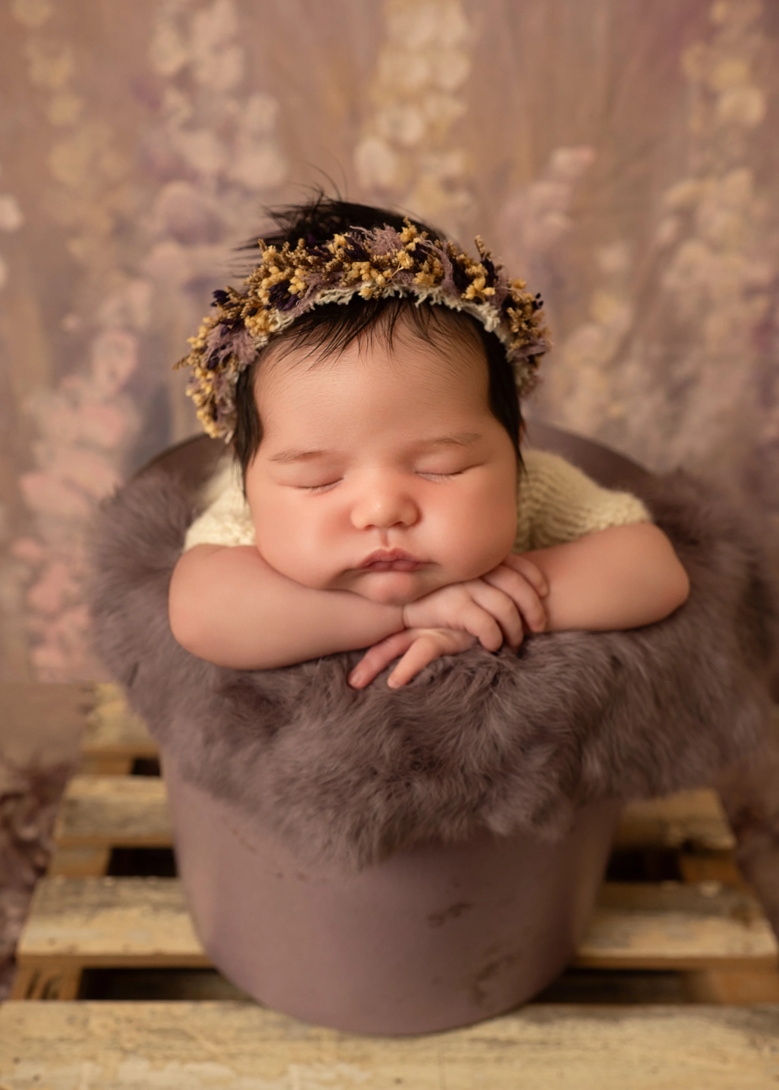 Baby sleeping in a knitted suit, styled in a purple bucket with her chin onher hands ontop of a dusty purple fur, with a dried floral headband, set against a floral background on top of a pallet.