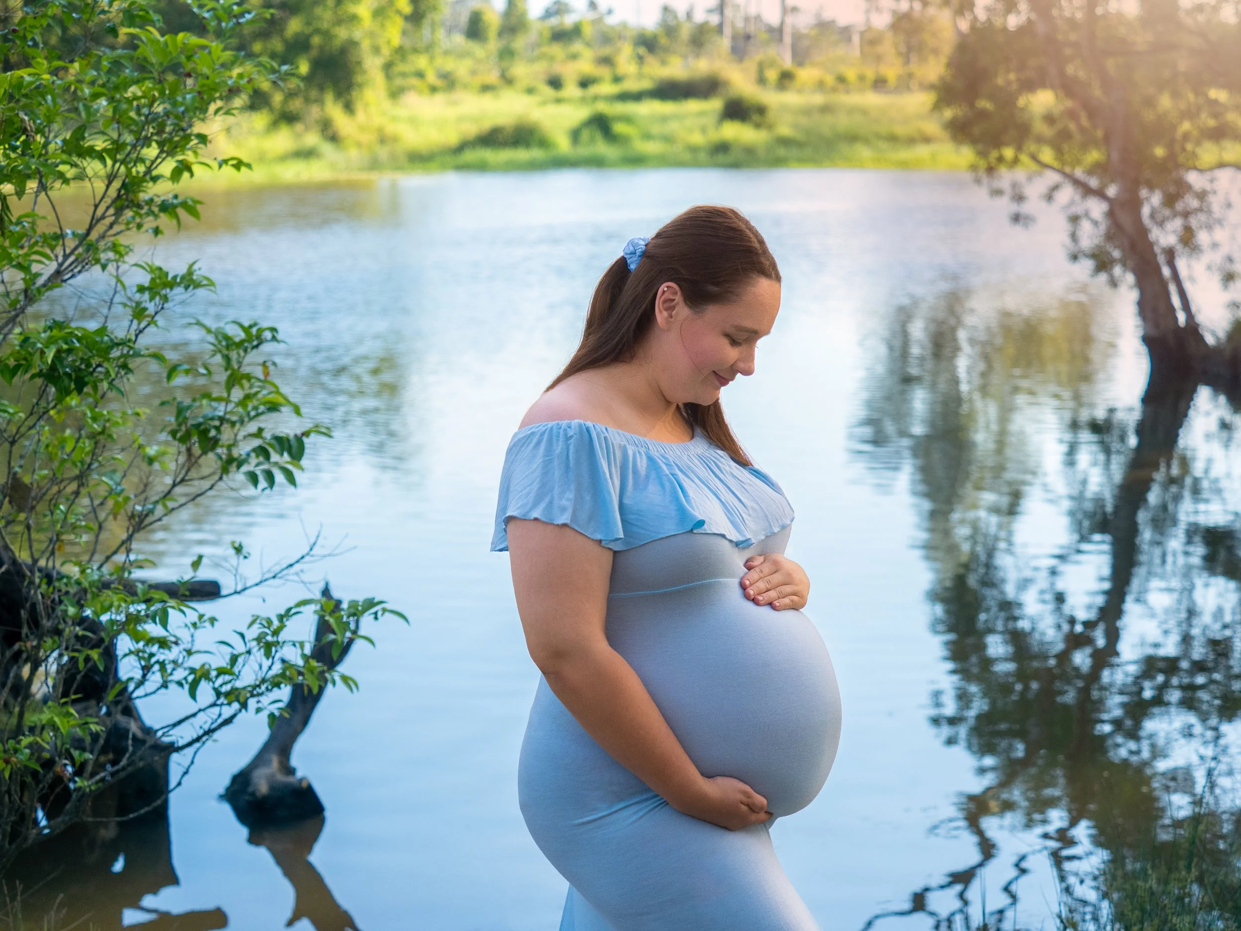 Pregnant mother in a pale blue dress cradling her baby bump near a Brisbane river.