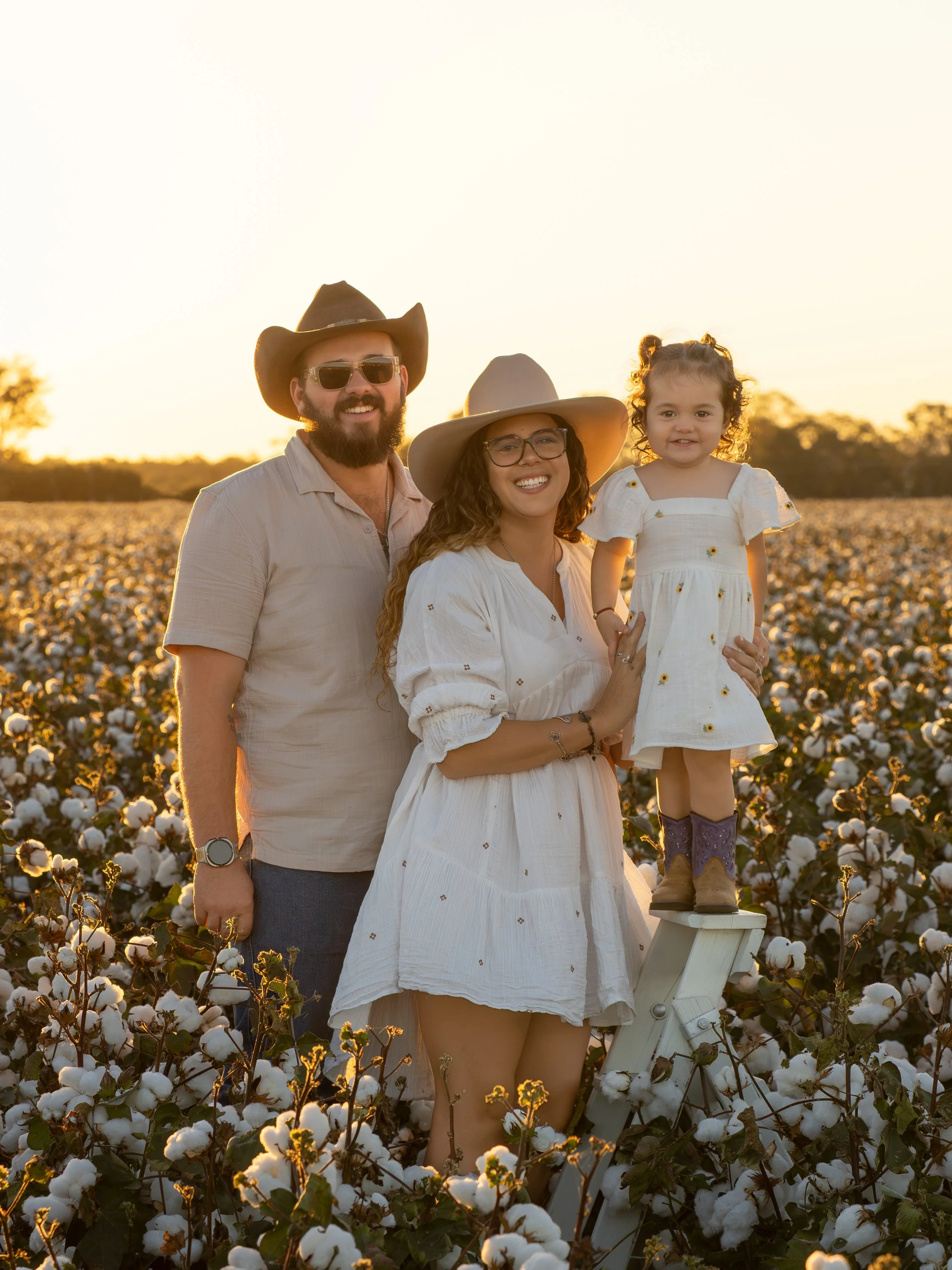 A smiling family of three, dressed in western attire, higging each other in the middle of a cotton field in the Lockyer Valley for a cotton sunset session.