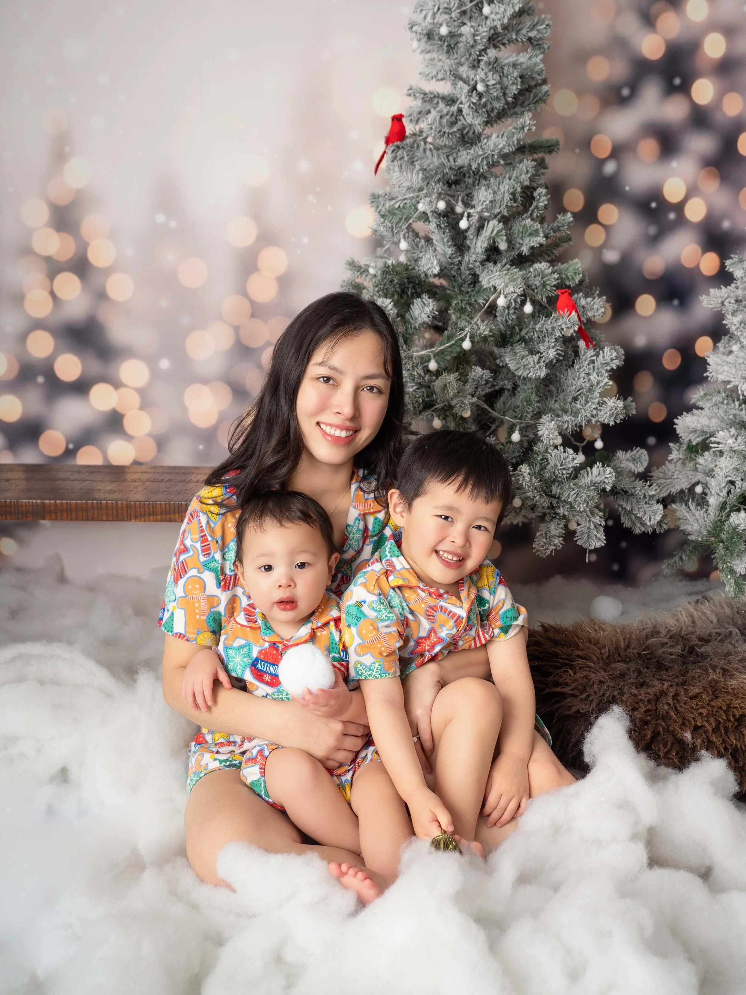 An Asian mother cuddles her two young sons with match Christmas pjs sitting in the snow in a winter wonderland Christmas set for mini photos