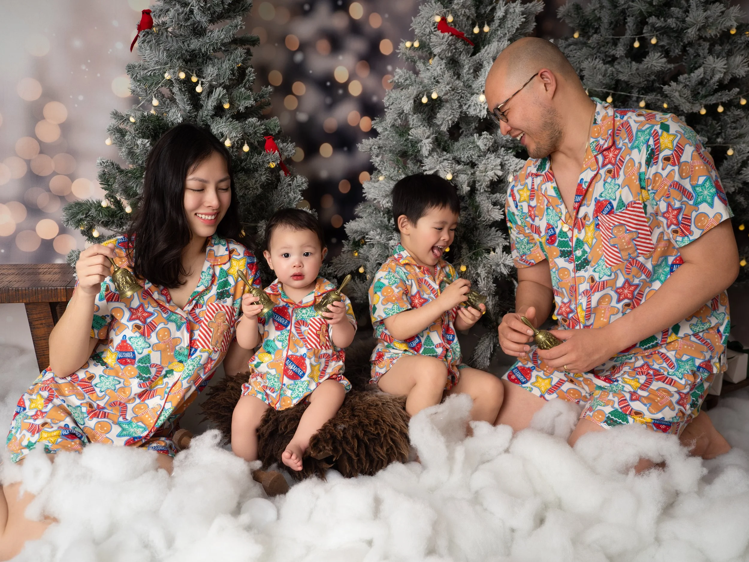 An Asian mother and father cheerfully ring small golden bells with theit two young boys in matching Christmas pajamas, nestled in the snow bank of the winter wonderland Christmas set