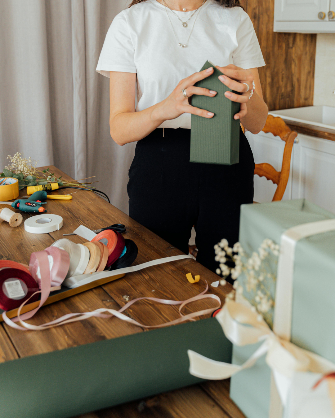 Person wrapping a green gift box in a room with a wooden table covered in wrapping supplies and floral decorations.