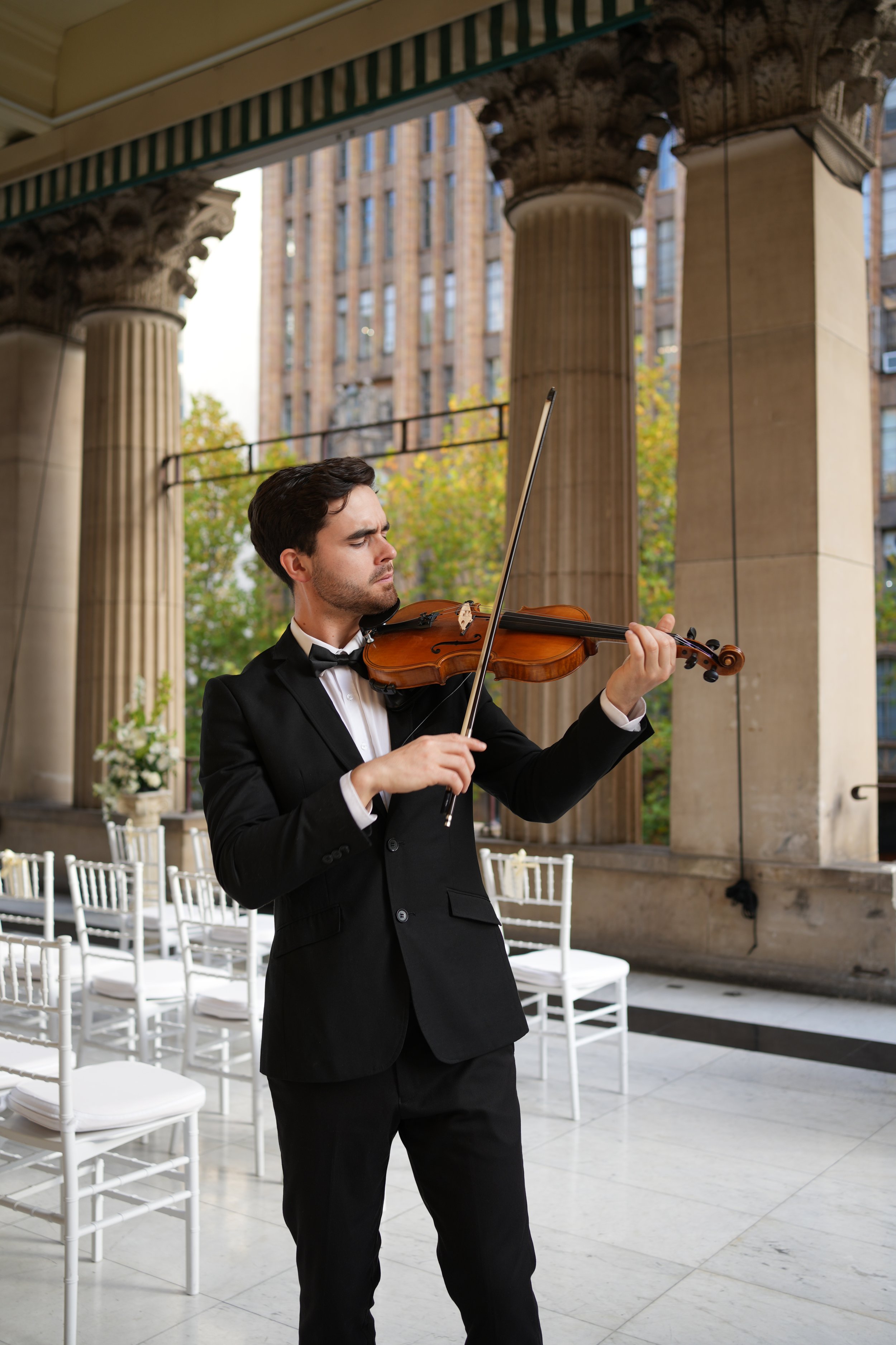 A young man dressed in a tuxedo playing the violin outdoors, with large columns and a cityscape with tall buildings in the background.