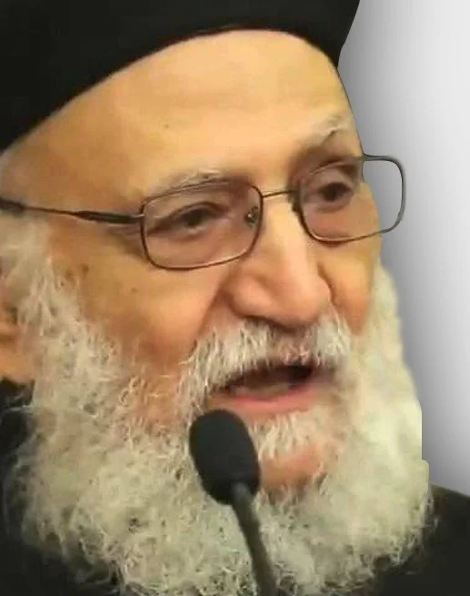 A close-up of an elderly man with a white beard, glasses, and a black turban, speaking into a microphone.
