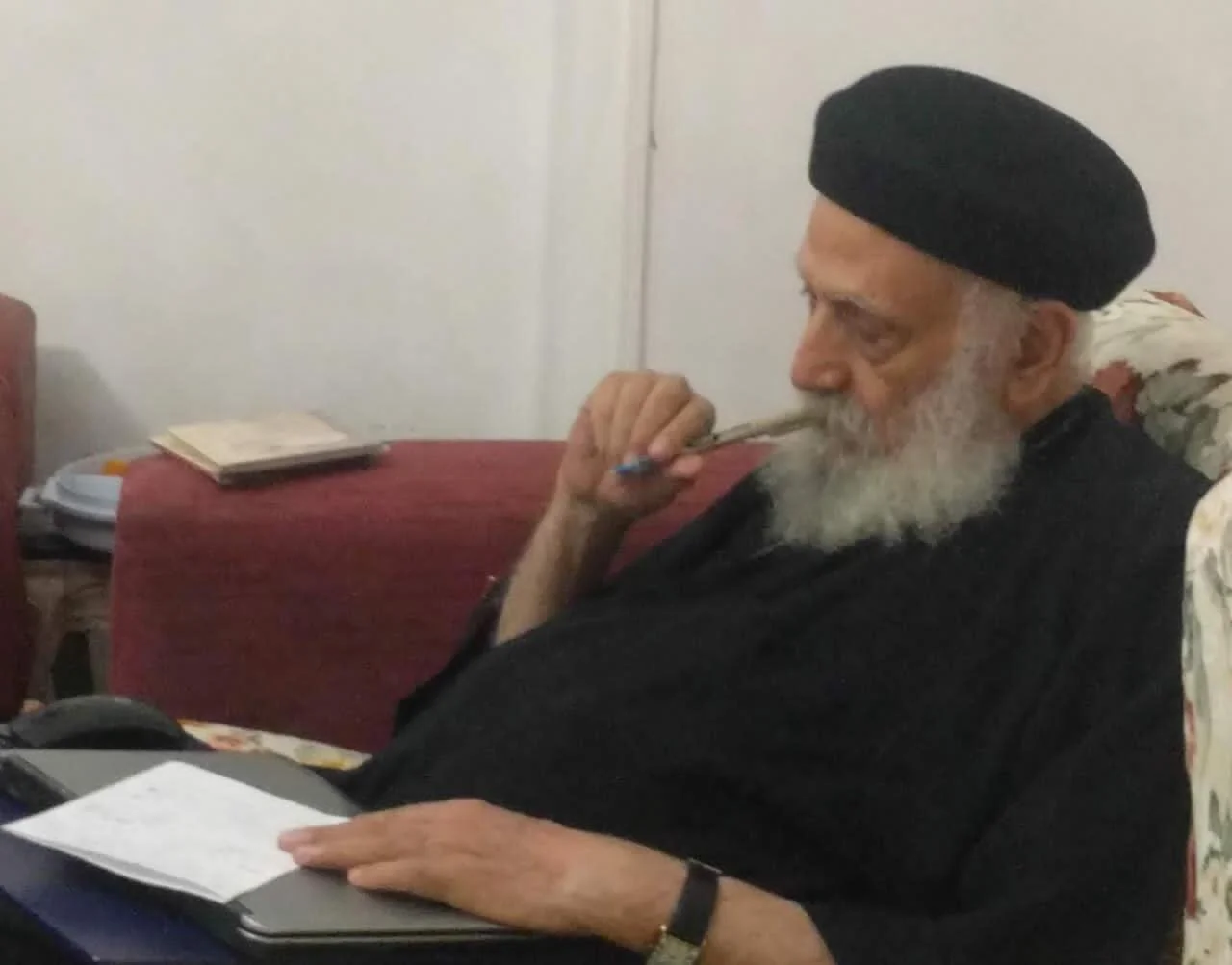 Abouna Tadros Y. Malaty sitting on a floral armchair, wearing a black hat and black clothing, holding a pen to his mouth while looking at a laptop.