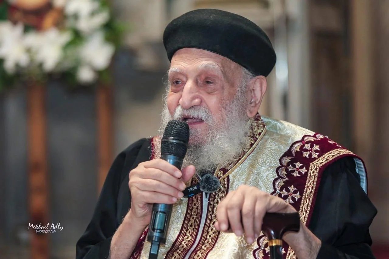 An elderly man with a white beard wearing traditional attire and a black hat speaking into a microphone at a formal event.