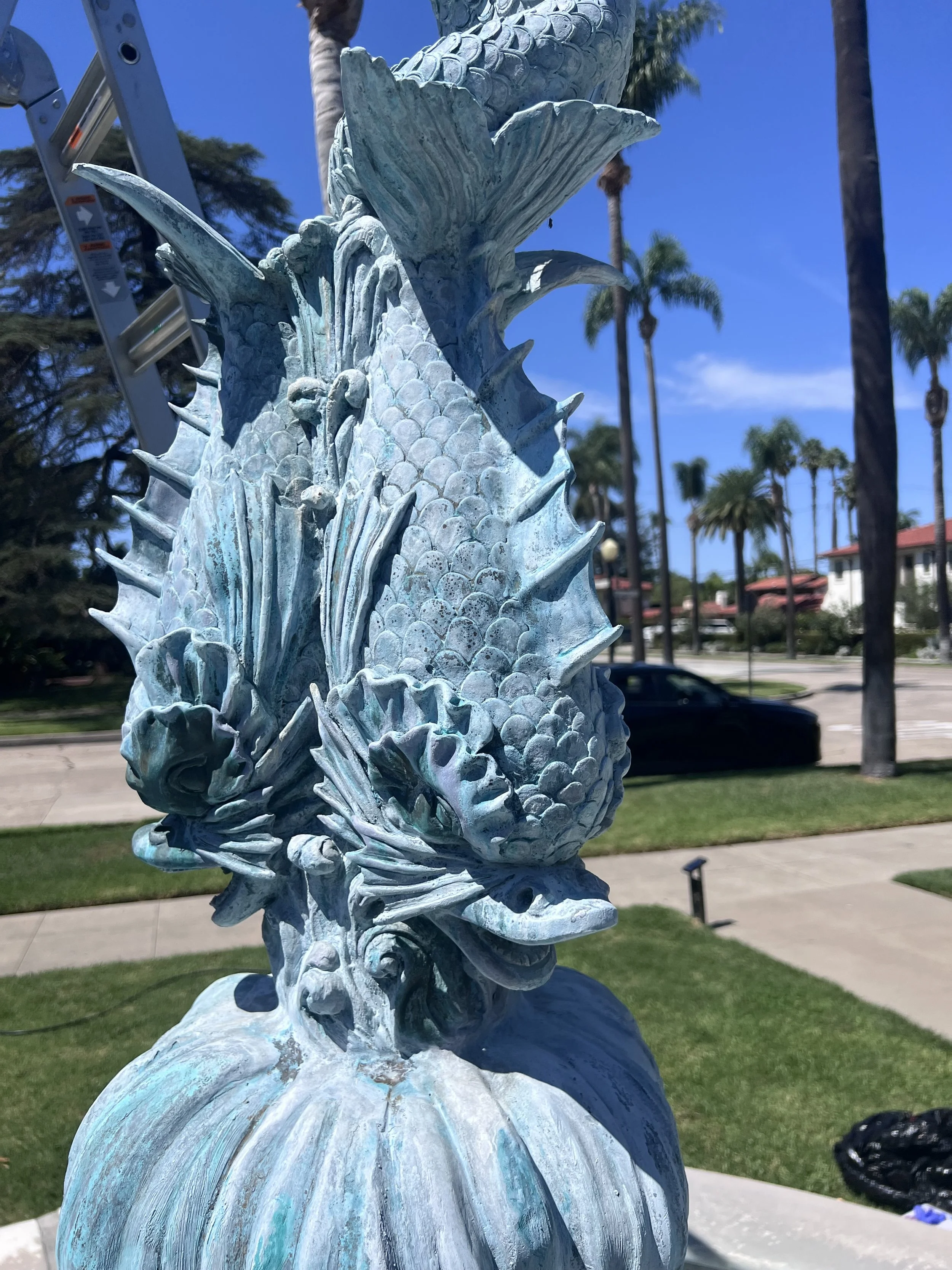 Close-up of a detailed metal sculpture of a seahorse with an oceanic patina, set outdoors with palm trees and a building in the background.