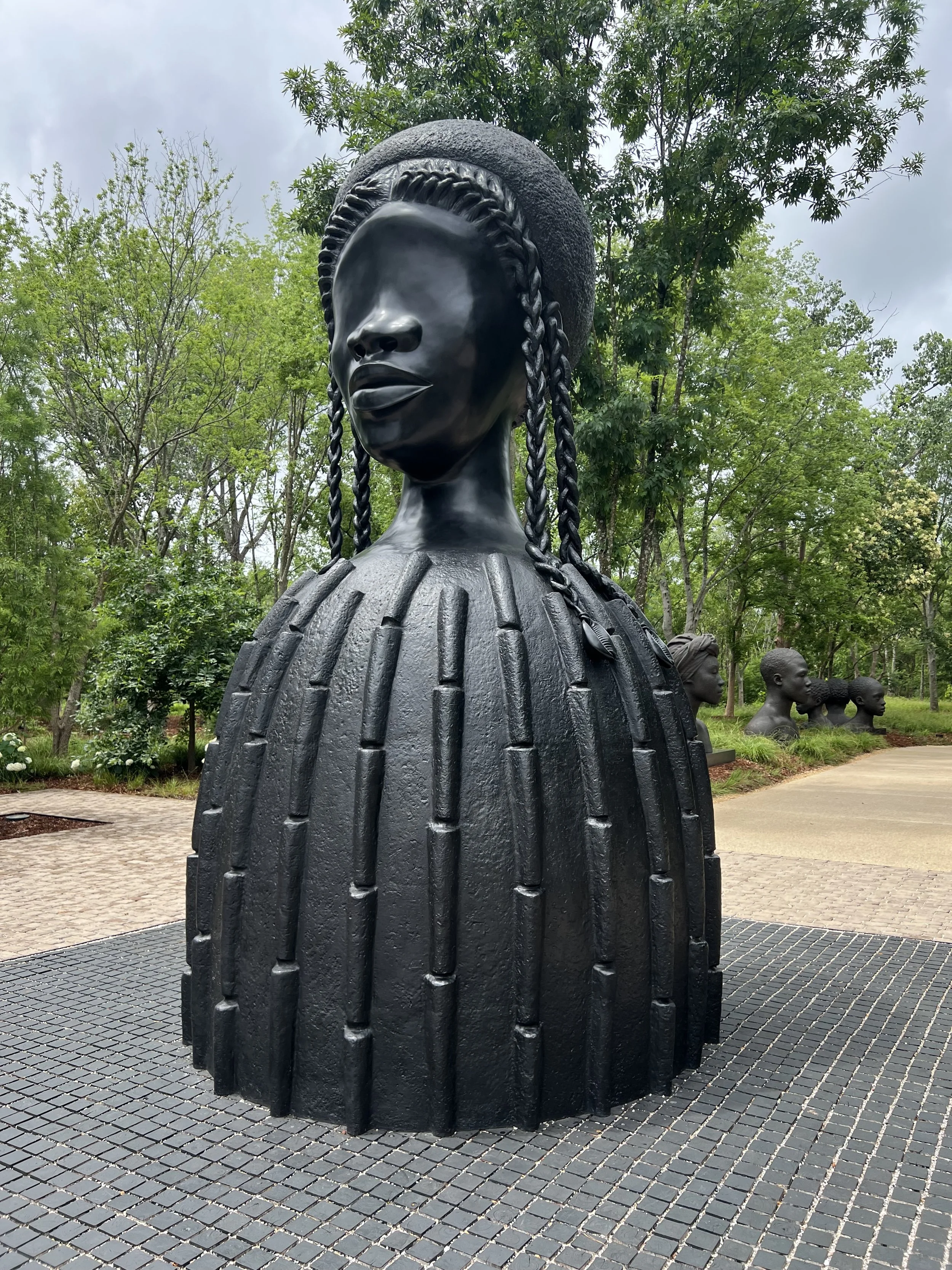 A large sculpture of a woman's head with braided hair, located outdoors among green trees and other similar sculptures in the background.