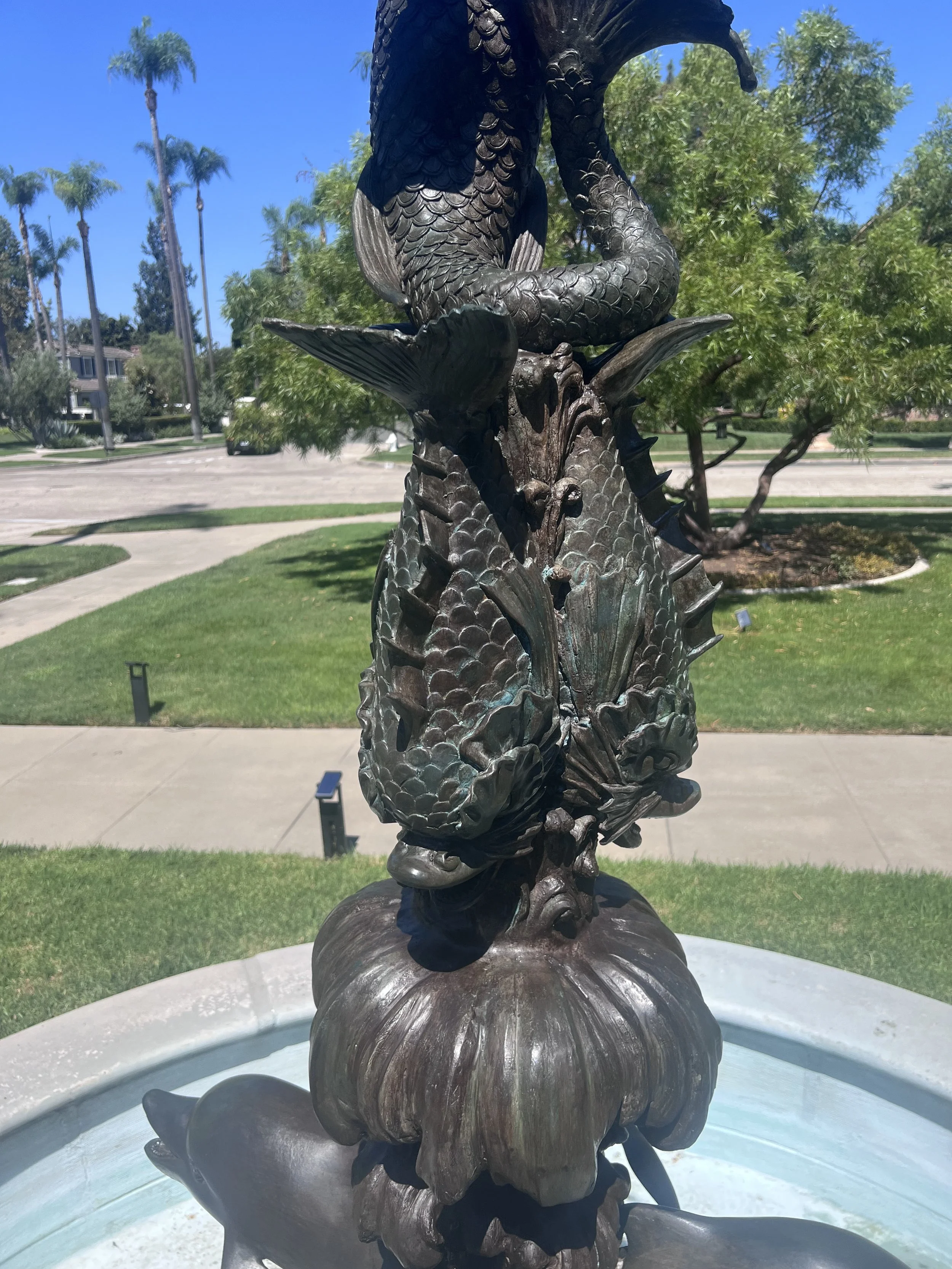 Bronze sculpture of a fish with a detailed, scale-like texture in an outdoor park setting with trees, palm trees, and a blue sky in the background.