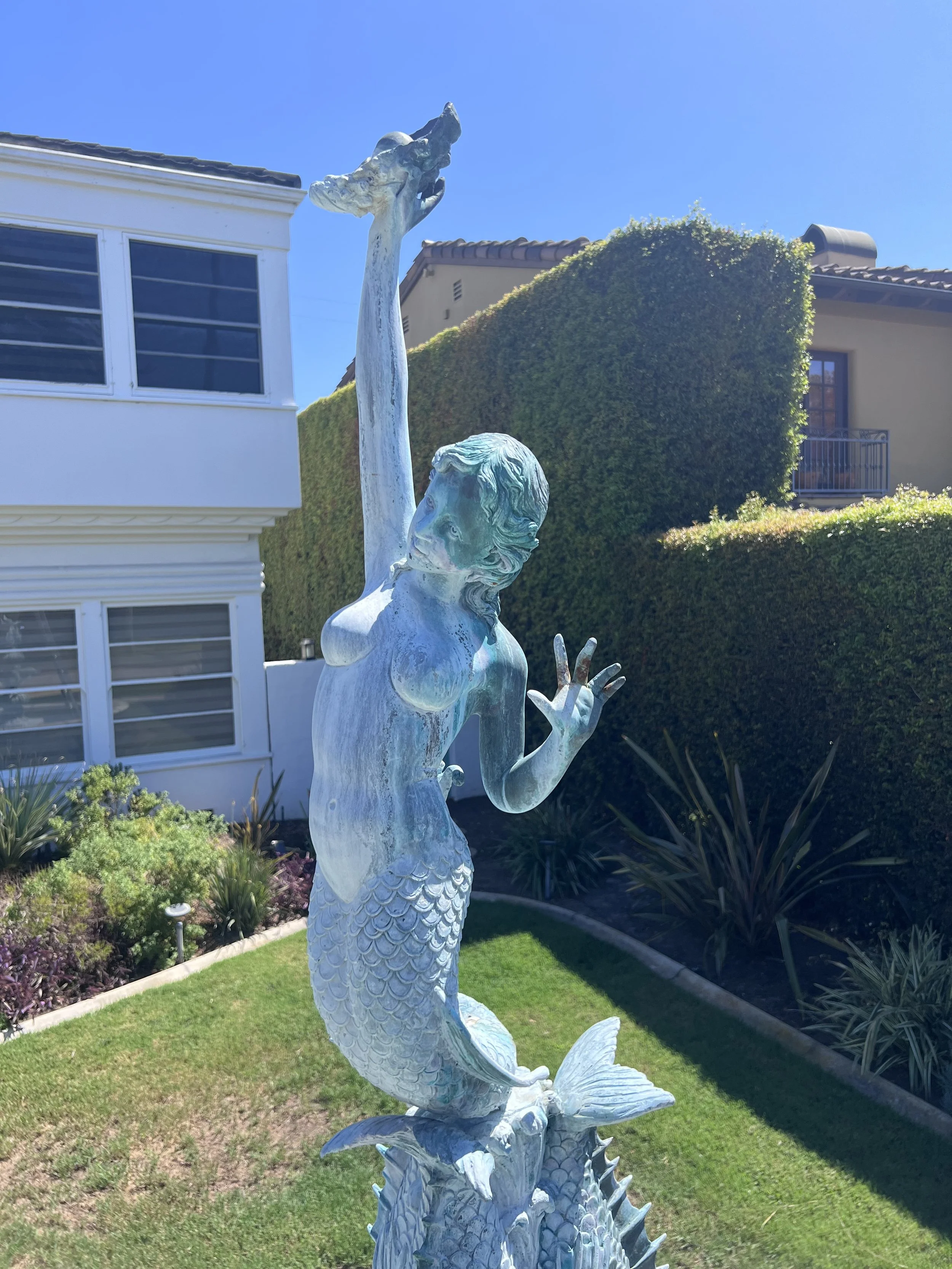 Statue of a mermaid with a fish tail and a human upper body, holding a large fish above her head, located in a well-maintained garden with green grass, shrubs, and a tall hedge, with residential buildings in the background and a bright blue sky.