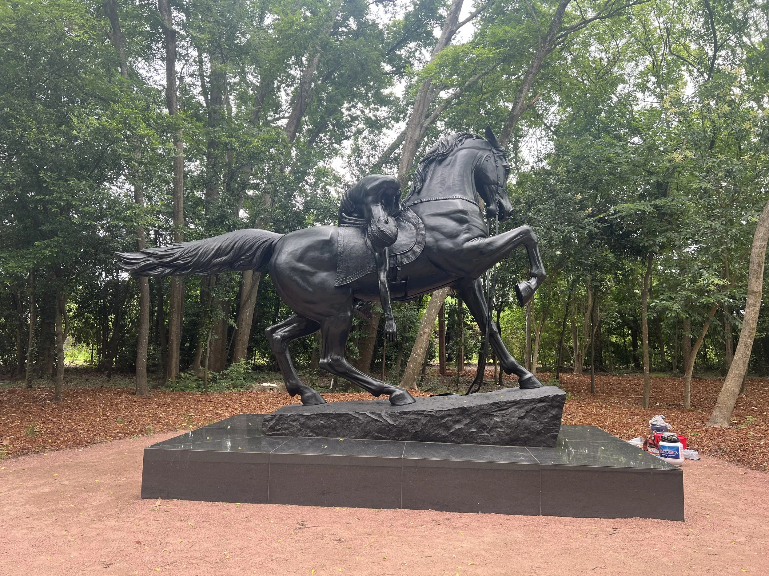 A bronze statue of a horse with a rider, located outdoors surrounded by trees and greenery.