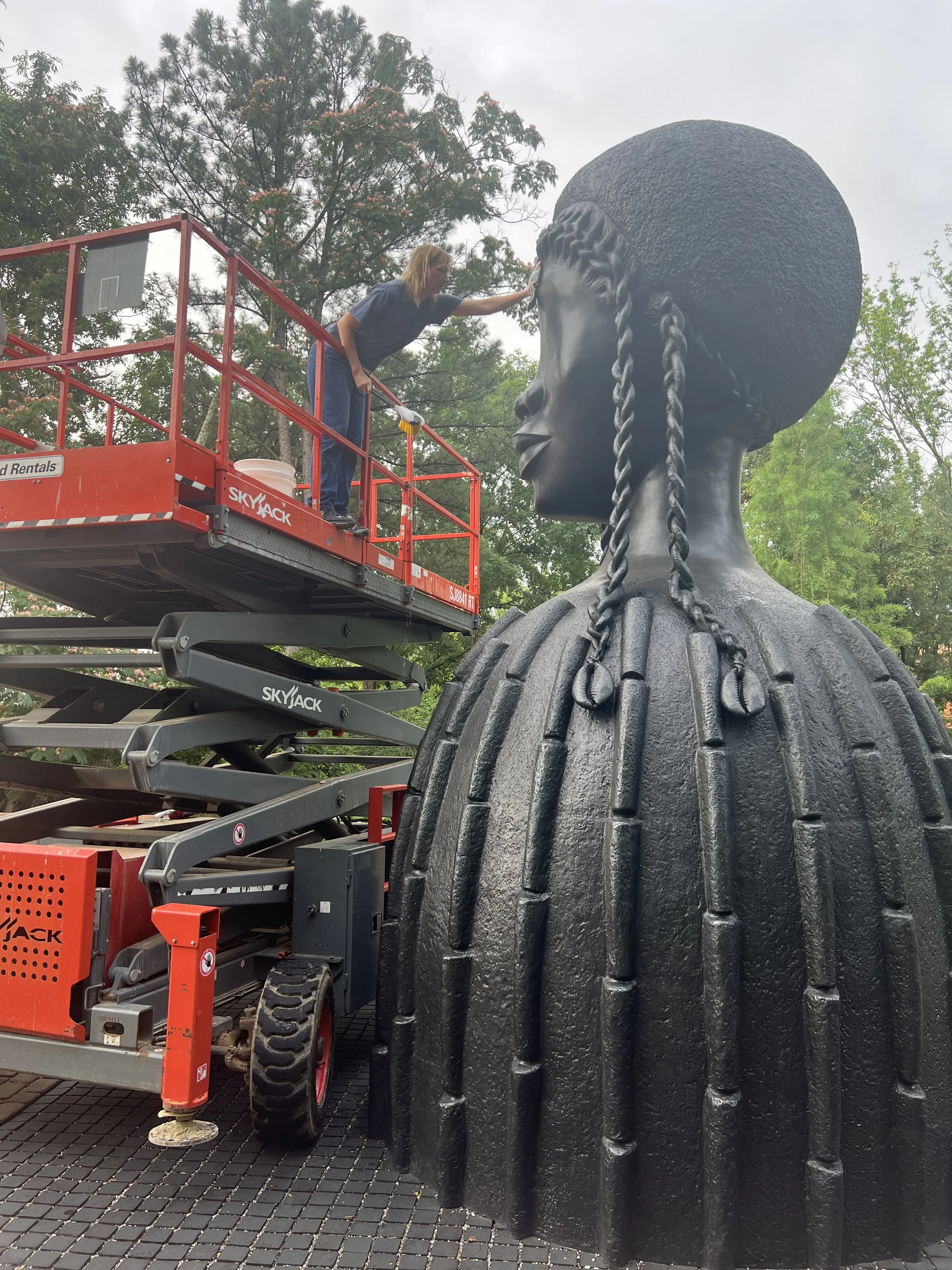A woman cleaning or restoring a large black bust sculpture of a woman's head and shoulders outdoors, on a lift platform with trees in the background.