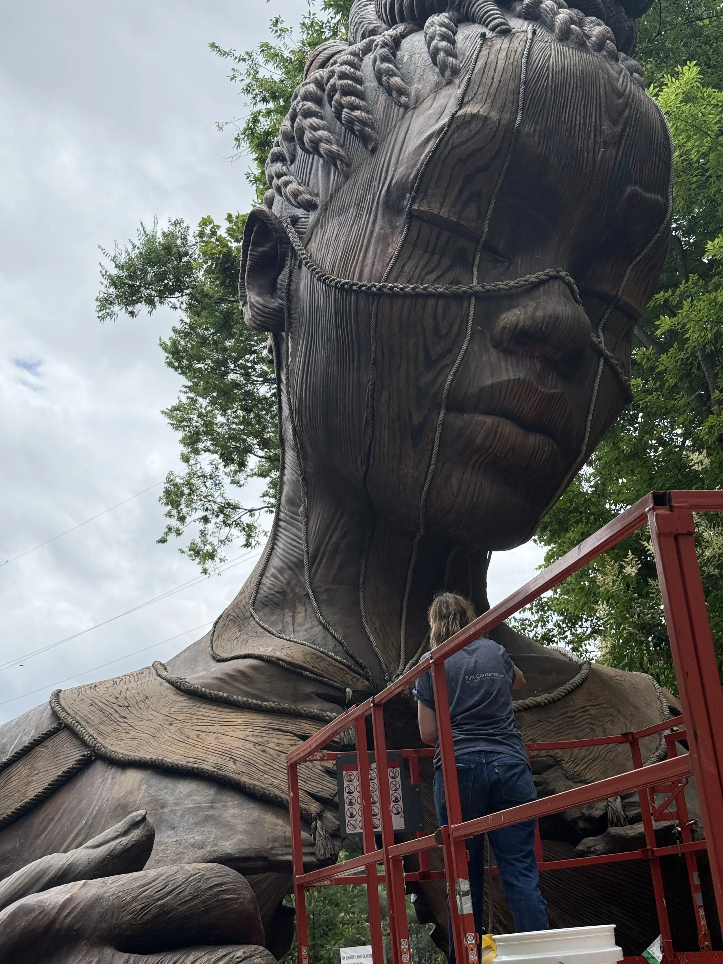 A worker on a red lift is in-painting a concrete sculpture 'Hallow' by Daniel Popper surrounded by green trees under a cloudy sky.