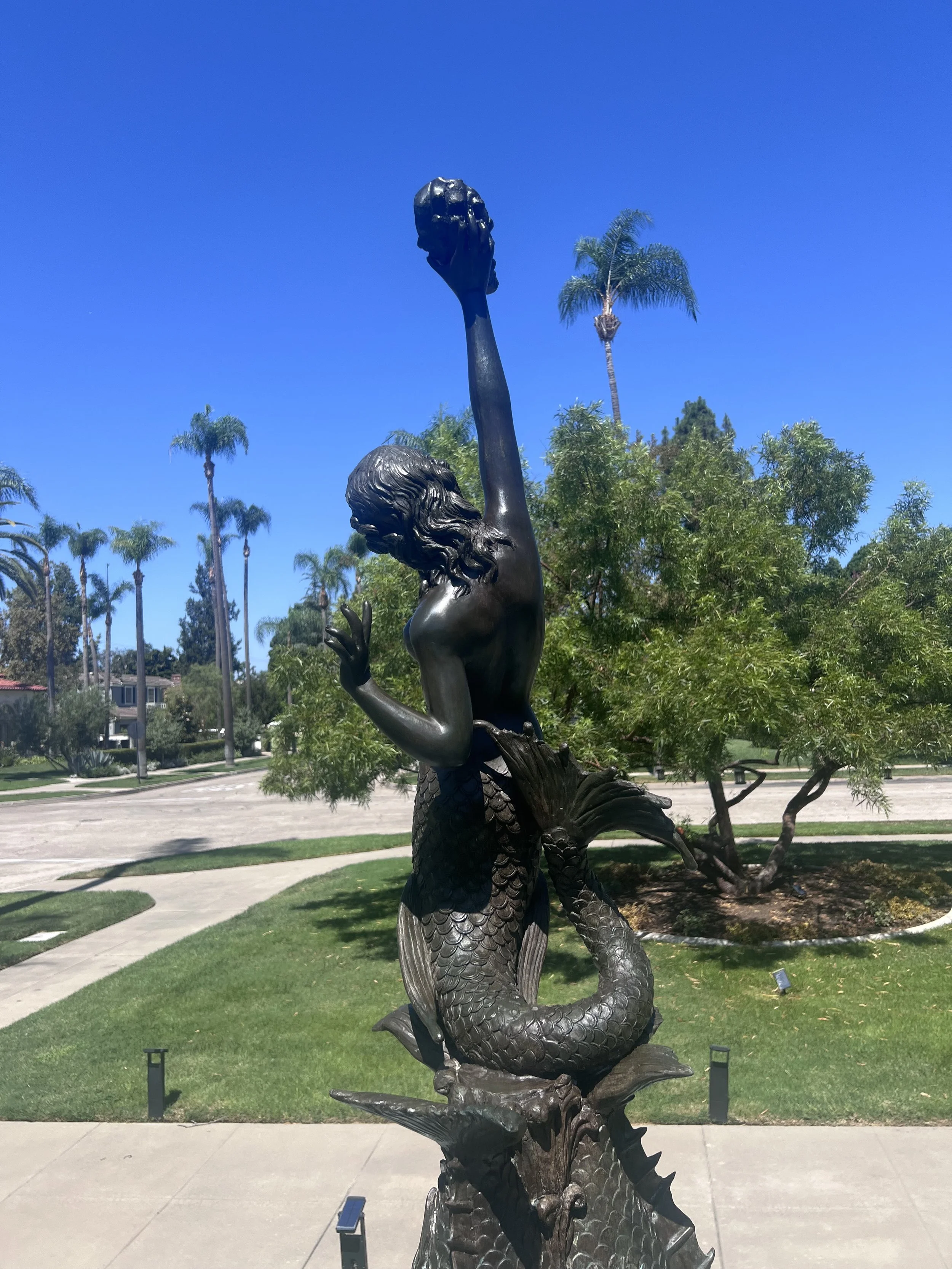 A bronze sculpture of a mermaid holding a fish above her head, located outdoors with palm trees and a clear blue sky in the background.