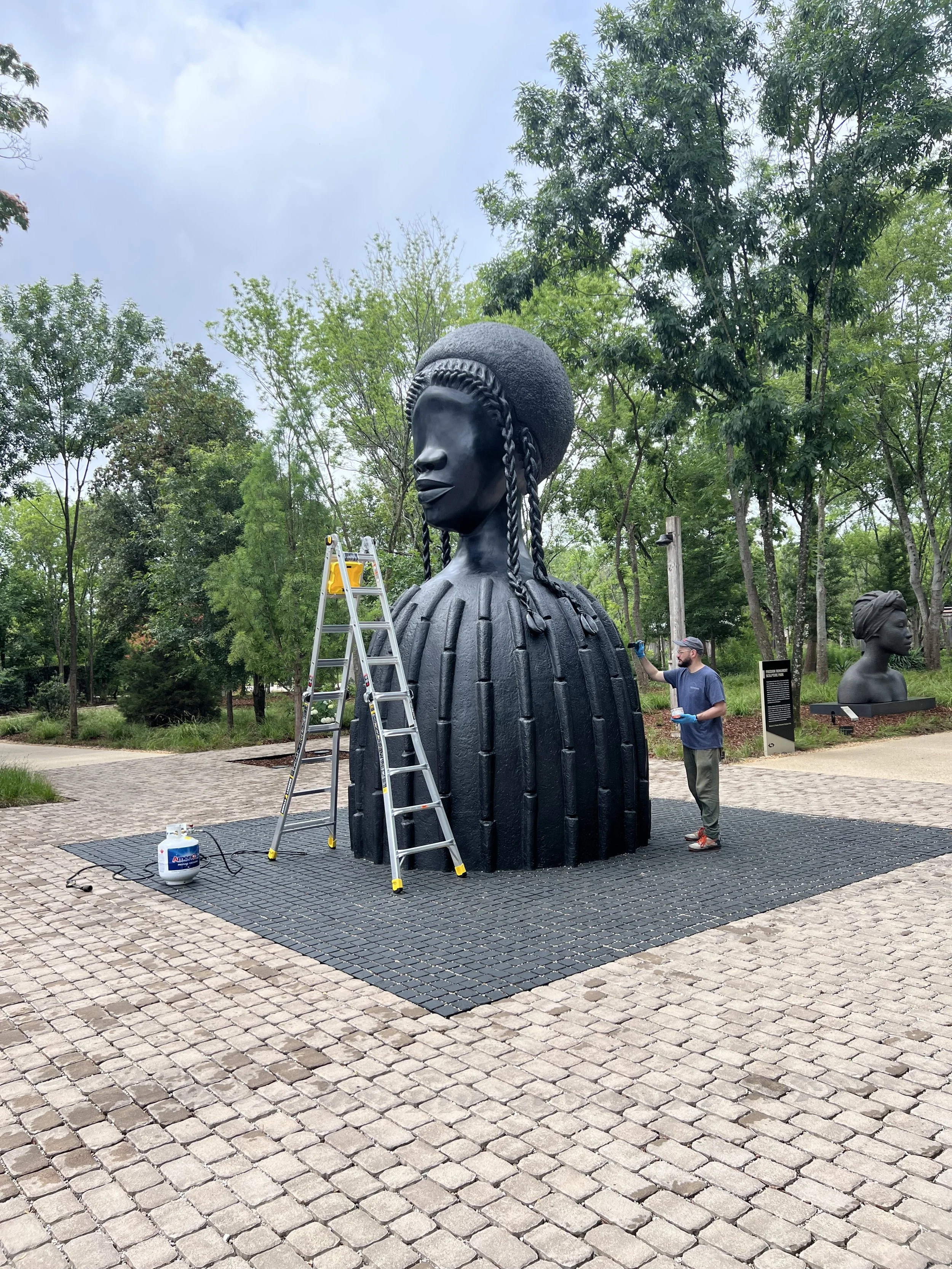 A large sculpture of a woman's head and shoulders, with braided hair and a rounded hairstyle, is shown outdoors. A man in gloves is working on the sculpture. A ladder is positioned next to it, and there is a propane tank nearby. The setting is a park