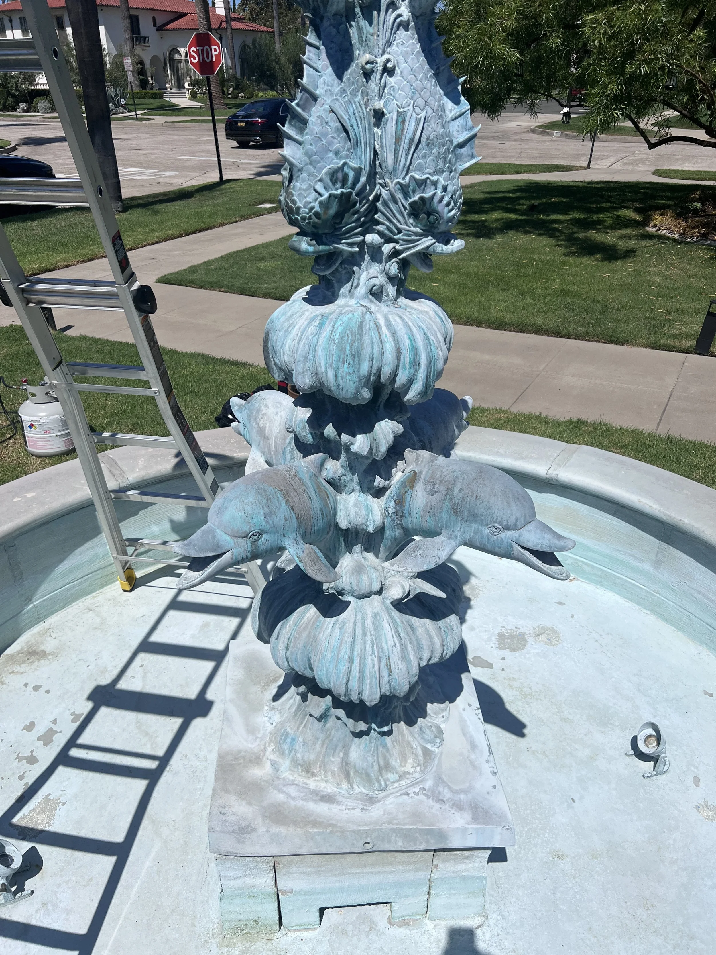 A decorative fountain featuring two dolphins in a circular basin, with a detailed sculpture of two seahorses and a large seashell on top, situated outdoors in a sunny area with houses, trees, and a stop sign visible in the background.