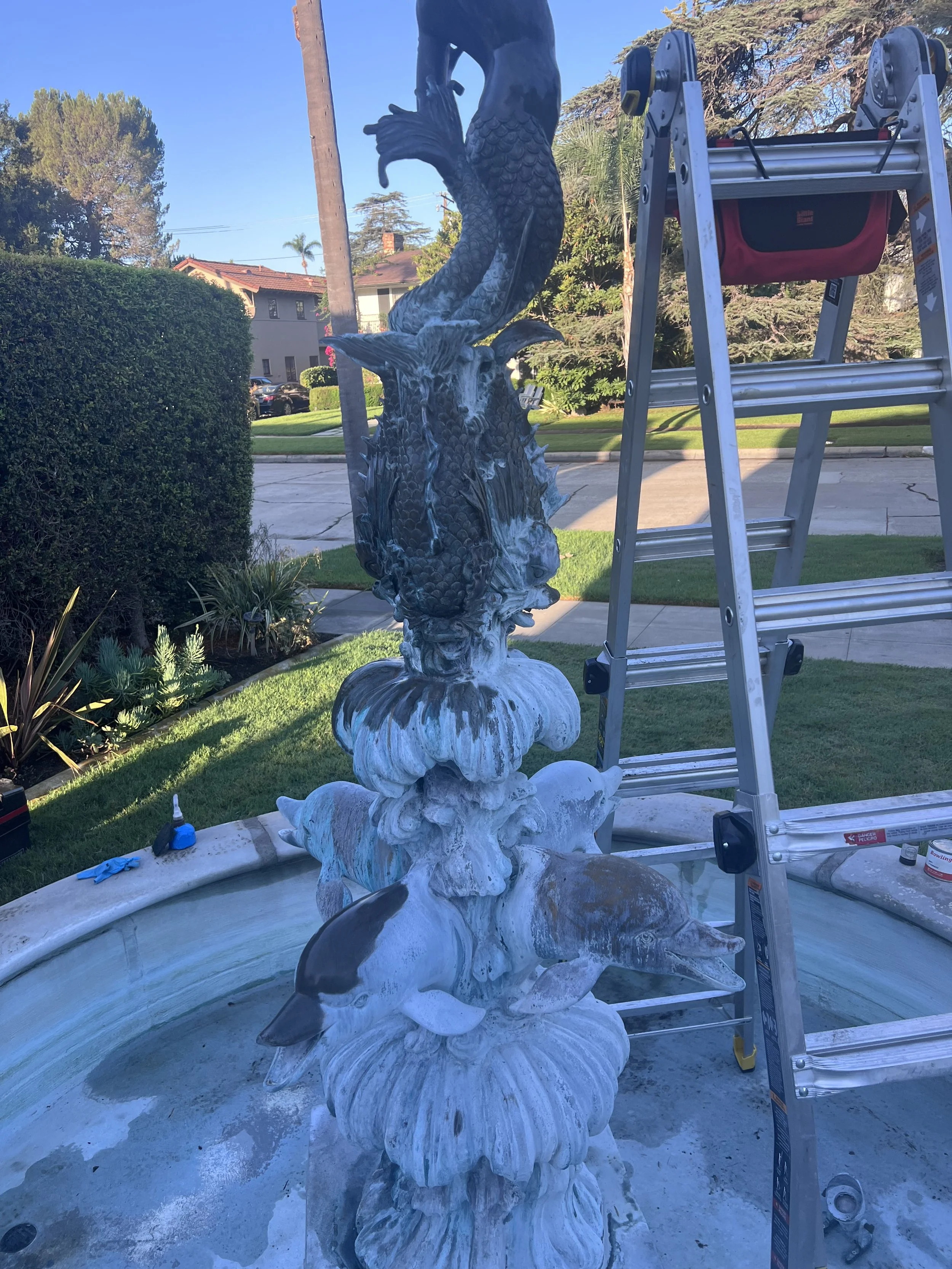 Outdoor scene showing a decorative fountain with fish and dragon figure sculptures, a ladder, and some tools on a concrete surface, with a background of trees, bushes, a street, and houses.
