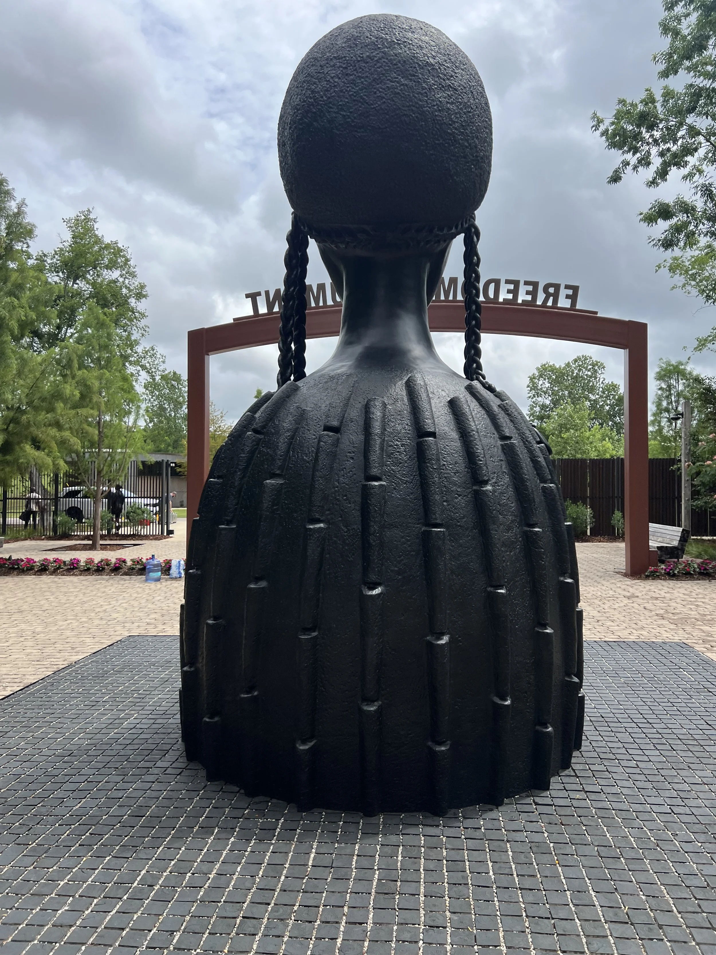 A large sculpture of a person from the back, with long braided hair, located outdoors in front of a sign that reads 'FREEDOM' in the background. 