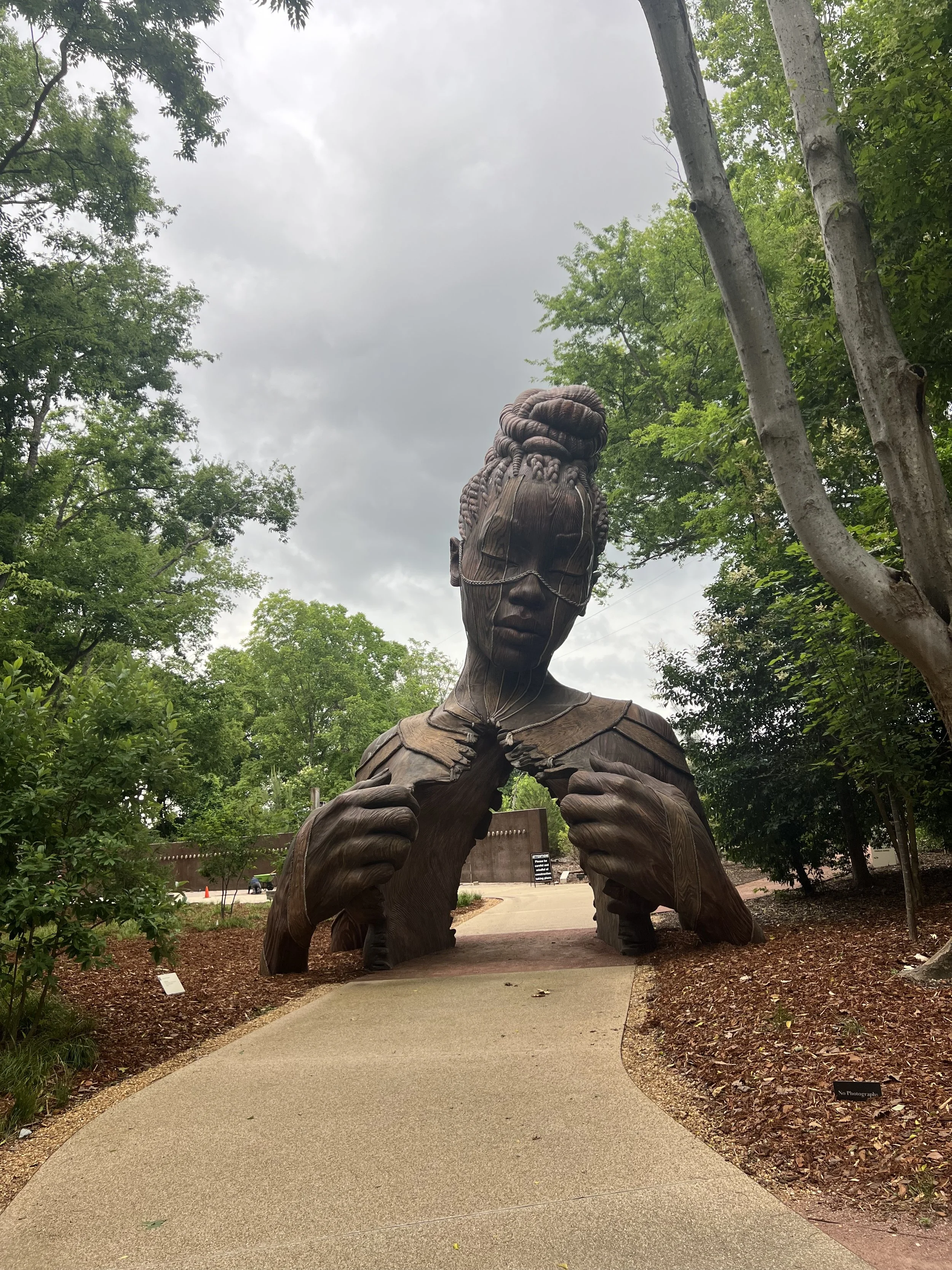 A large wooden sculpture of a person with their head down and hands pulling their chest open, located outdoors among trees and greenery.