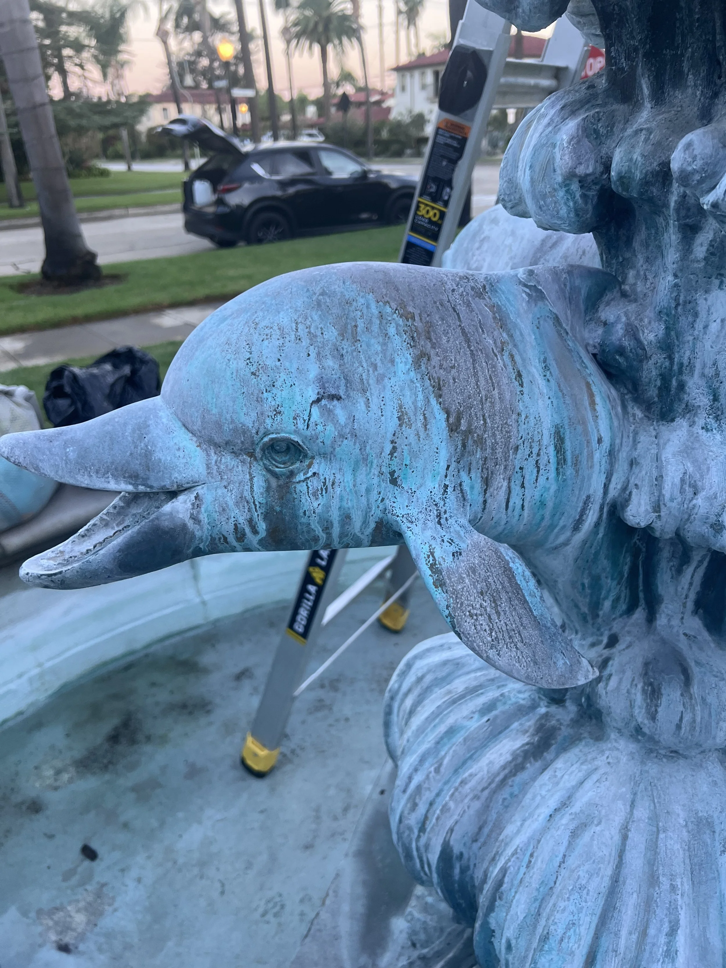 Close-up of a dolphin fountain sculpture with a teal patina, outdoors on a sunny day, with a black SUV with its trunk open, a ladder, and houses in the background.