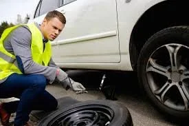 Man in a yellow safety vest changing a flat tire on a white vehicle.
