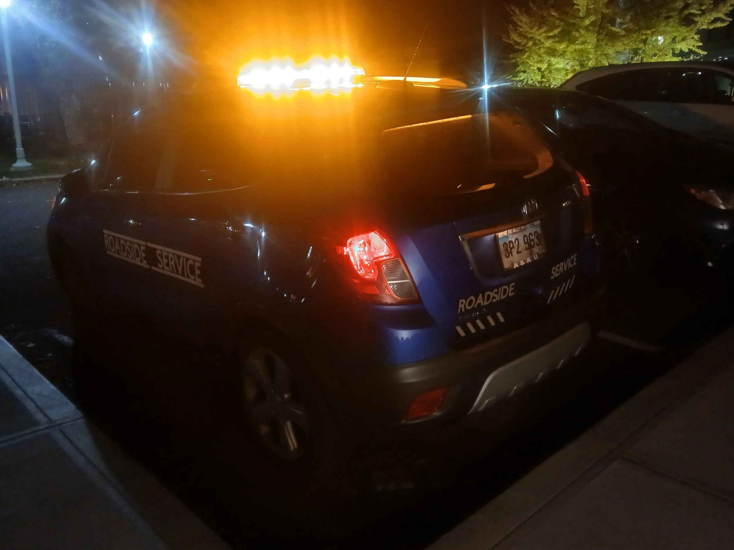 Dark blue vehicle with 'Roadside Service' stickers, parked on a street at night with flashing orange and red emergency lights on top.