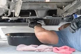 A person lying on their back, reaching under a car with an oil pan or container to catch or drain fluid, possibly changing the oil.